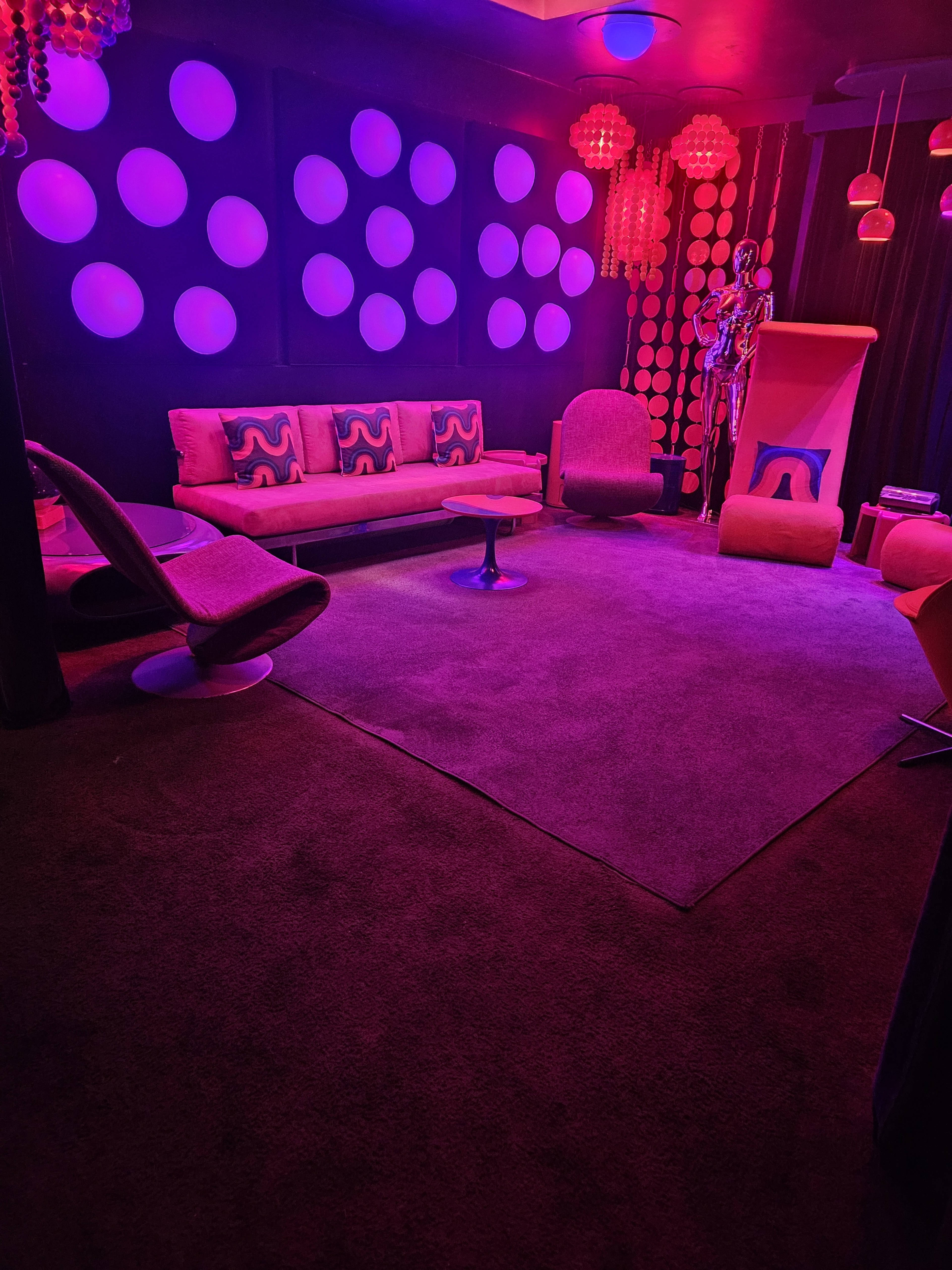 The image shows a retro-styled lounge area with circular wall decorations, pink lighting, and modern furniture, including a sofa and armchairs.