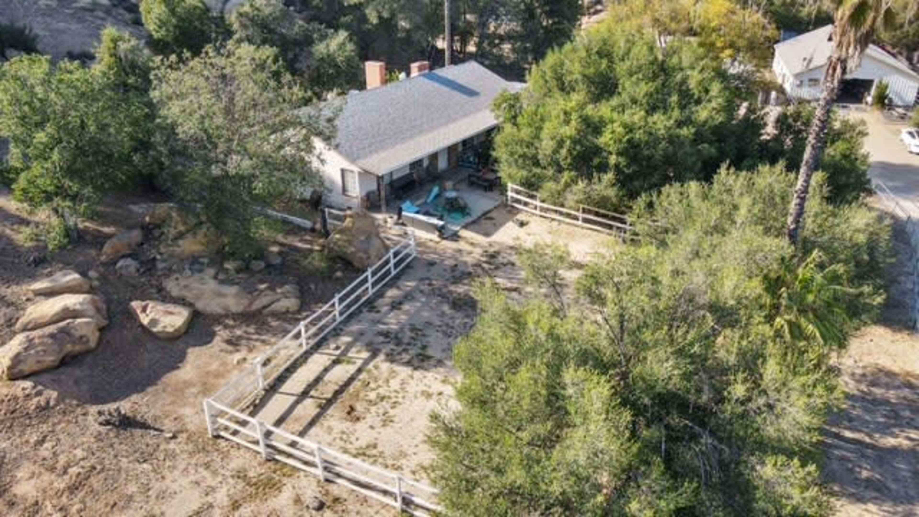 The image shows a single-story house surrounded by trees and a white fence, with large rocks and a dirt area nearby.