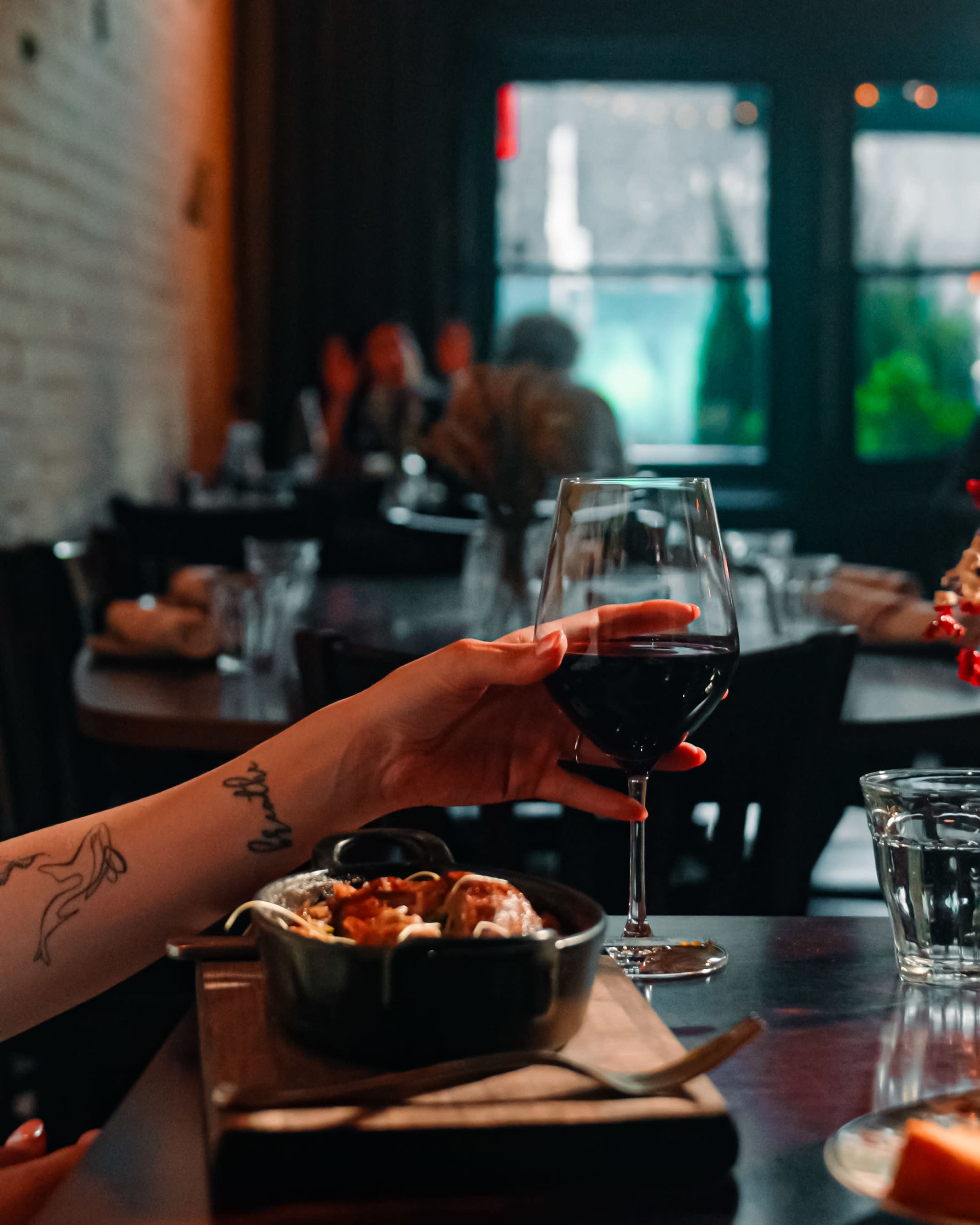 A hand holding a glass of red wine reaches toward a small dish on a wooden table in a dimly lit restaurant.