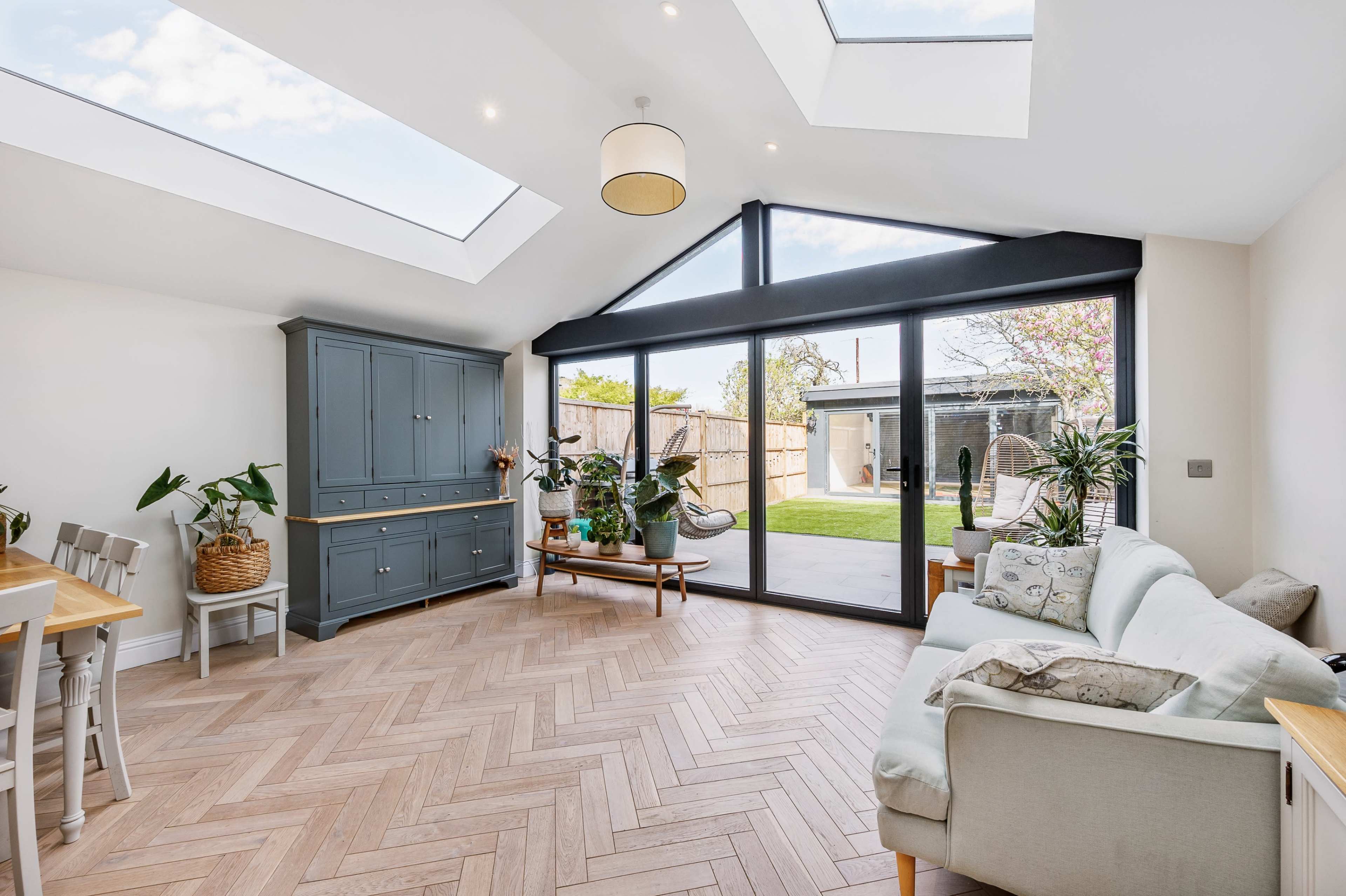 A bright, modern living space with large windows, a herringbone-patterned wooden floor, and comfortable furniture.