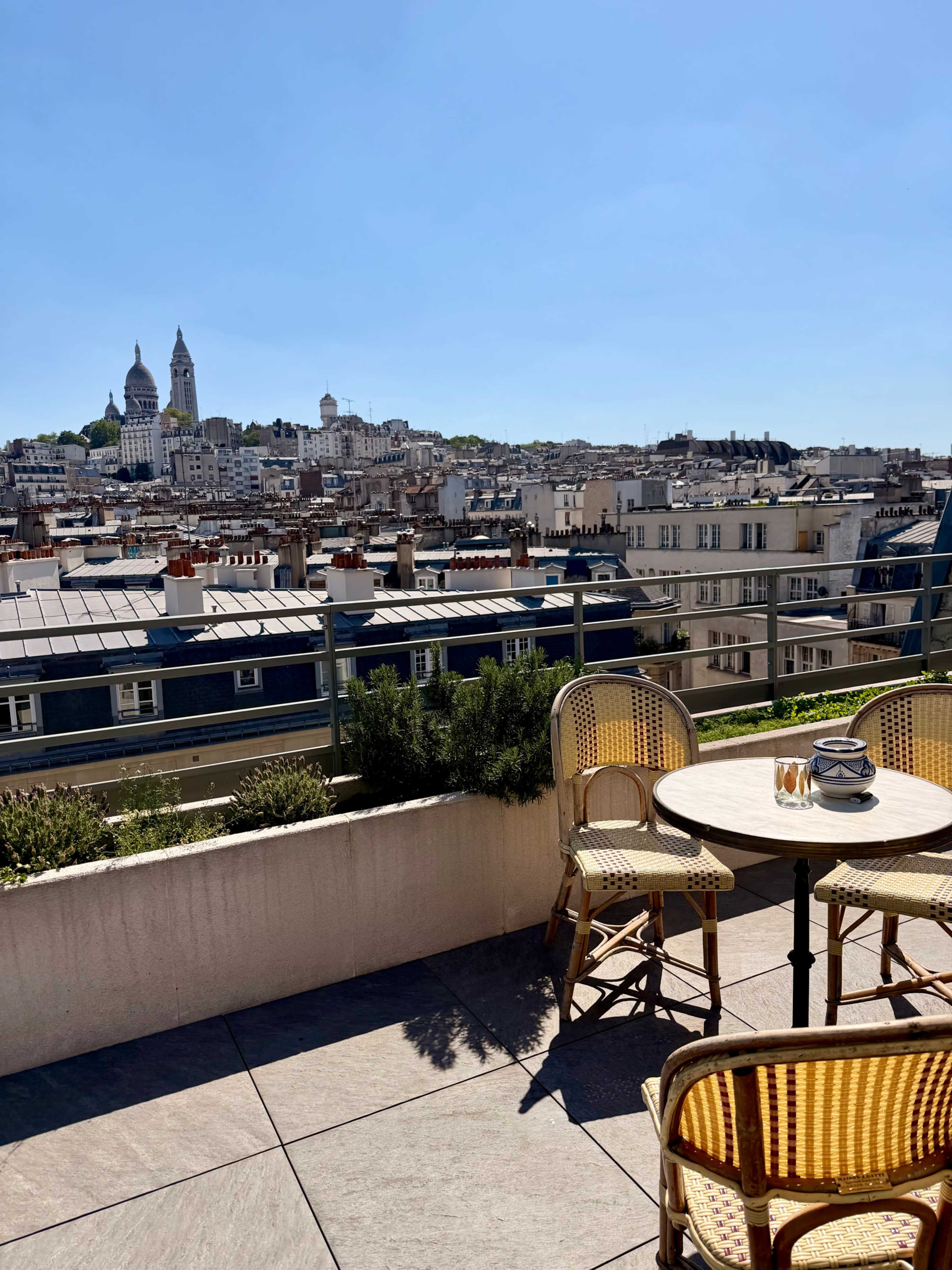 The image shows a rooftop balcony with a small round table and two wicker chairs, overlooking a cityscape featuring buildings and a church in the distance.