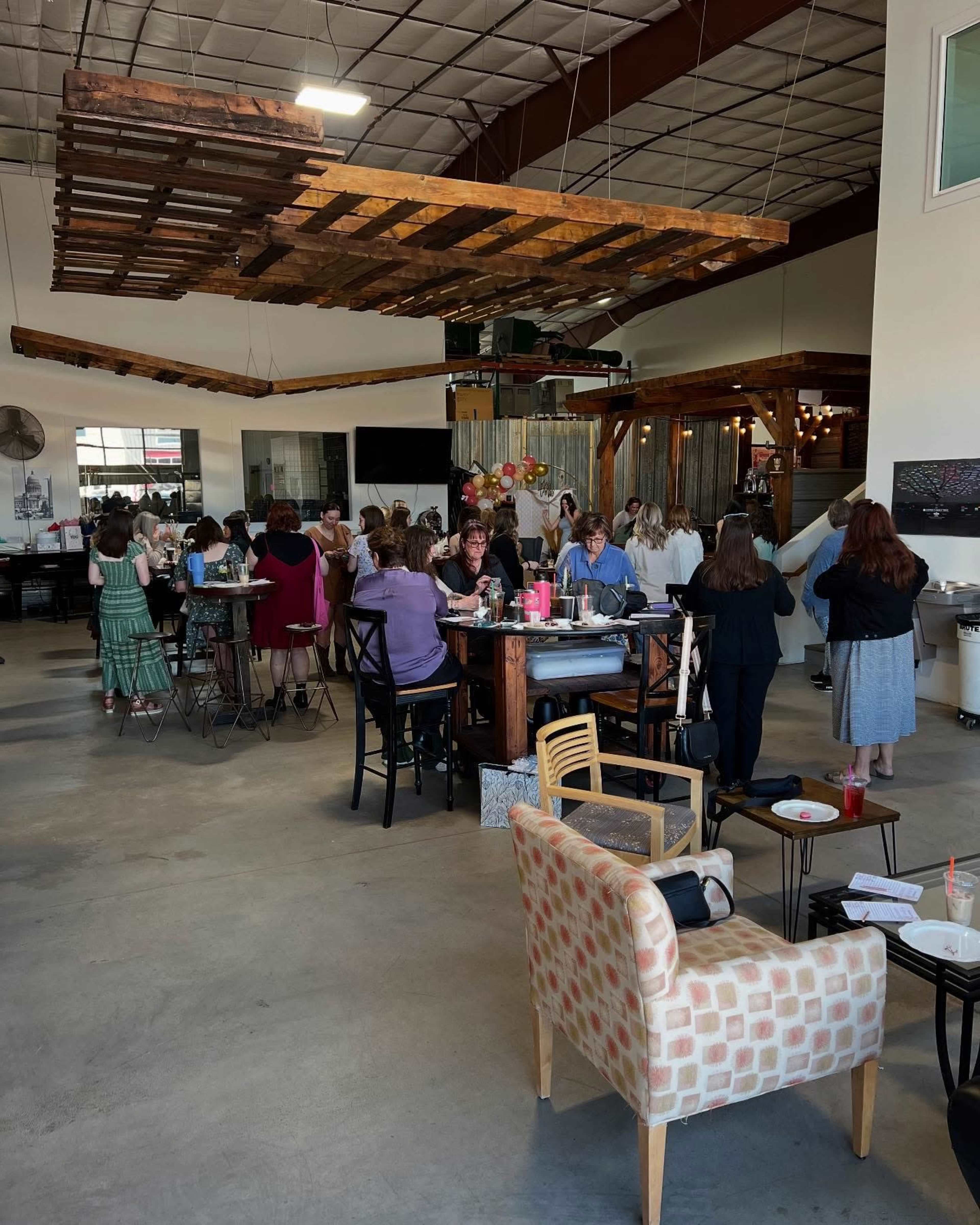 A bustling indoor space features a large group of people seated at tables, engaging in activities, under a wooden pallet installation hanging from the ceiling.