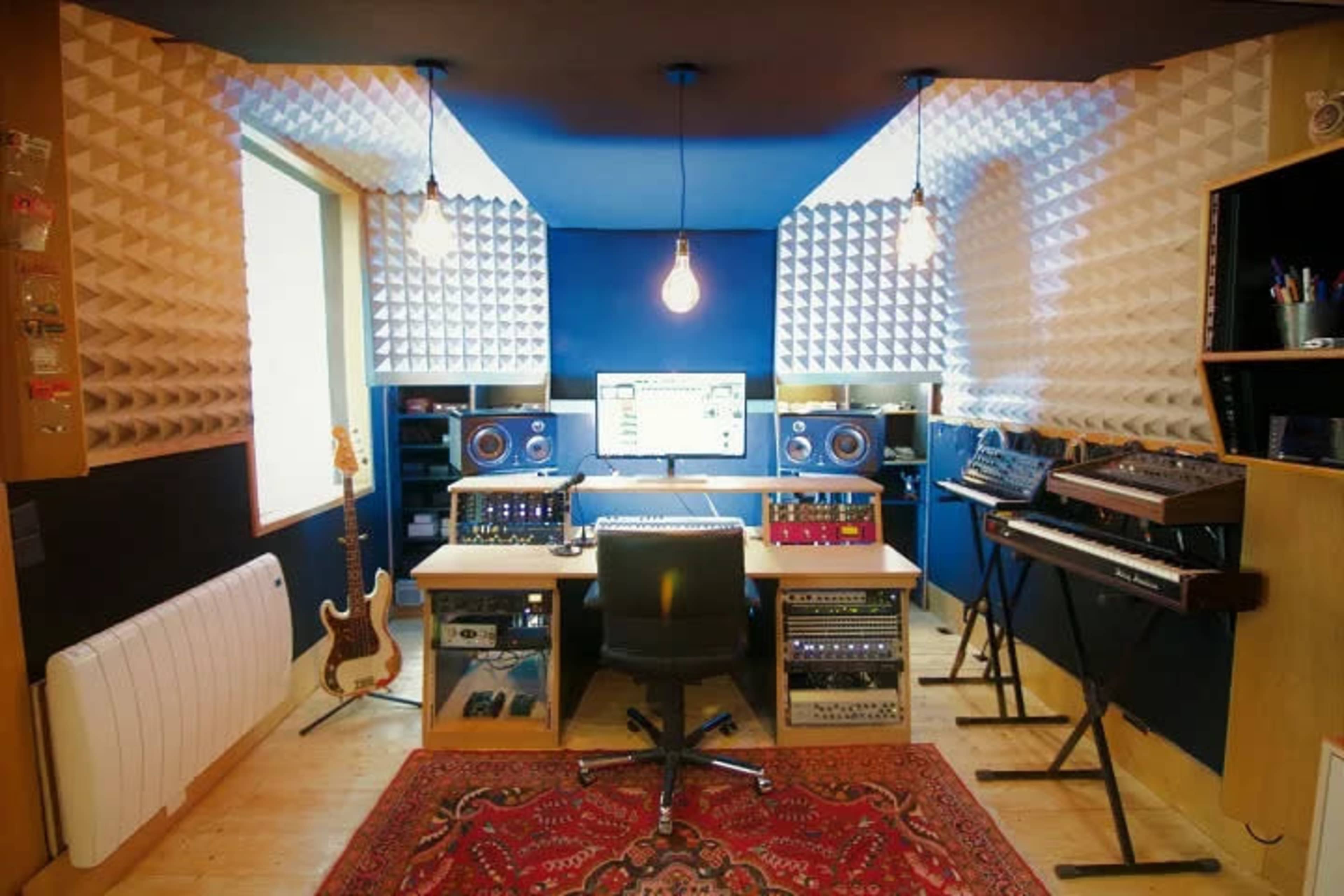 The image shows a well-equipped music studio featuring a central desk with audio equipment, a large computer monitor, and soundproofing panels on the walls.