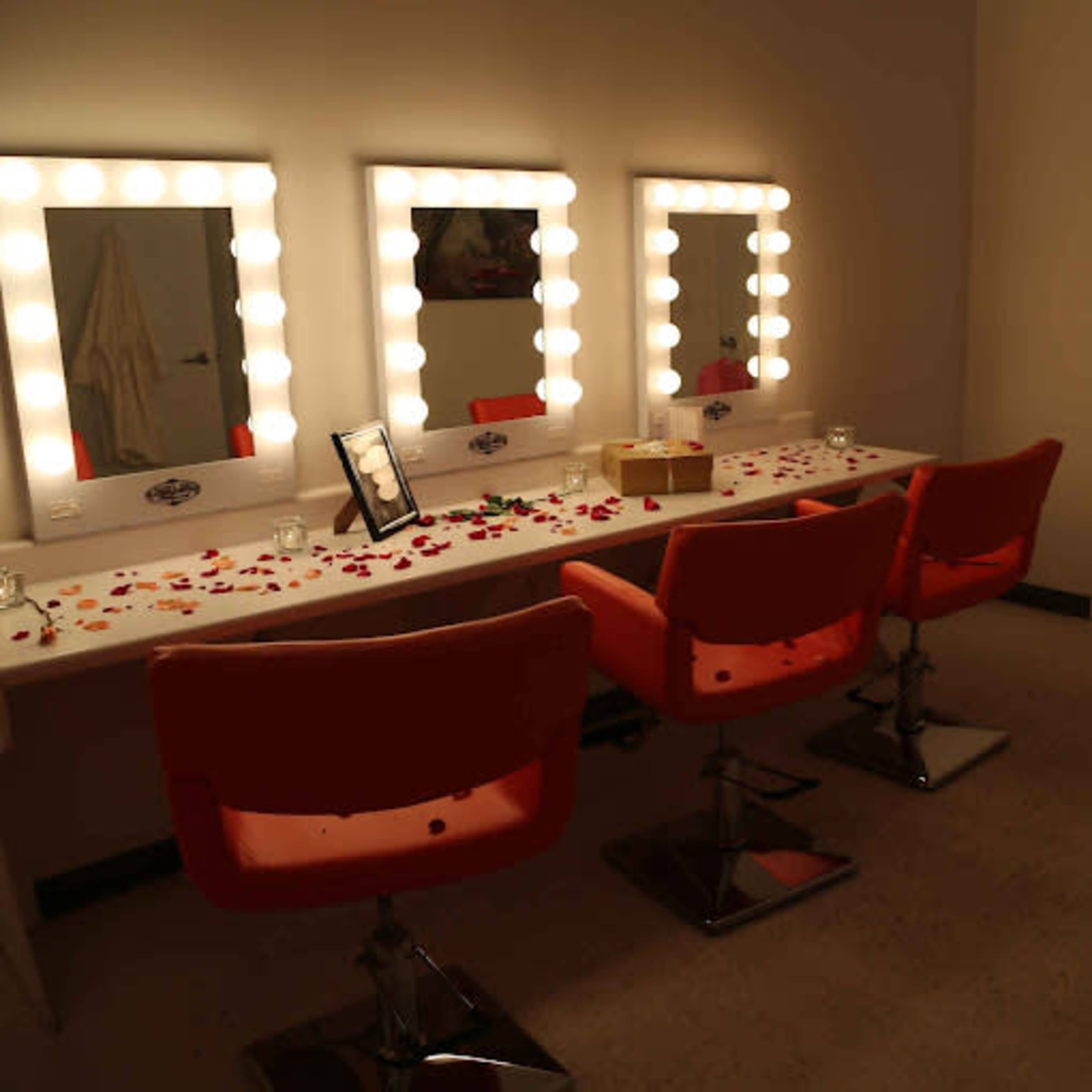 A makeup station with three illuminated mirrors, orange chairs, and a table decorated with rose petals and small items.