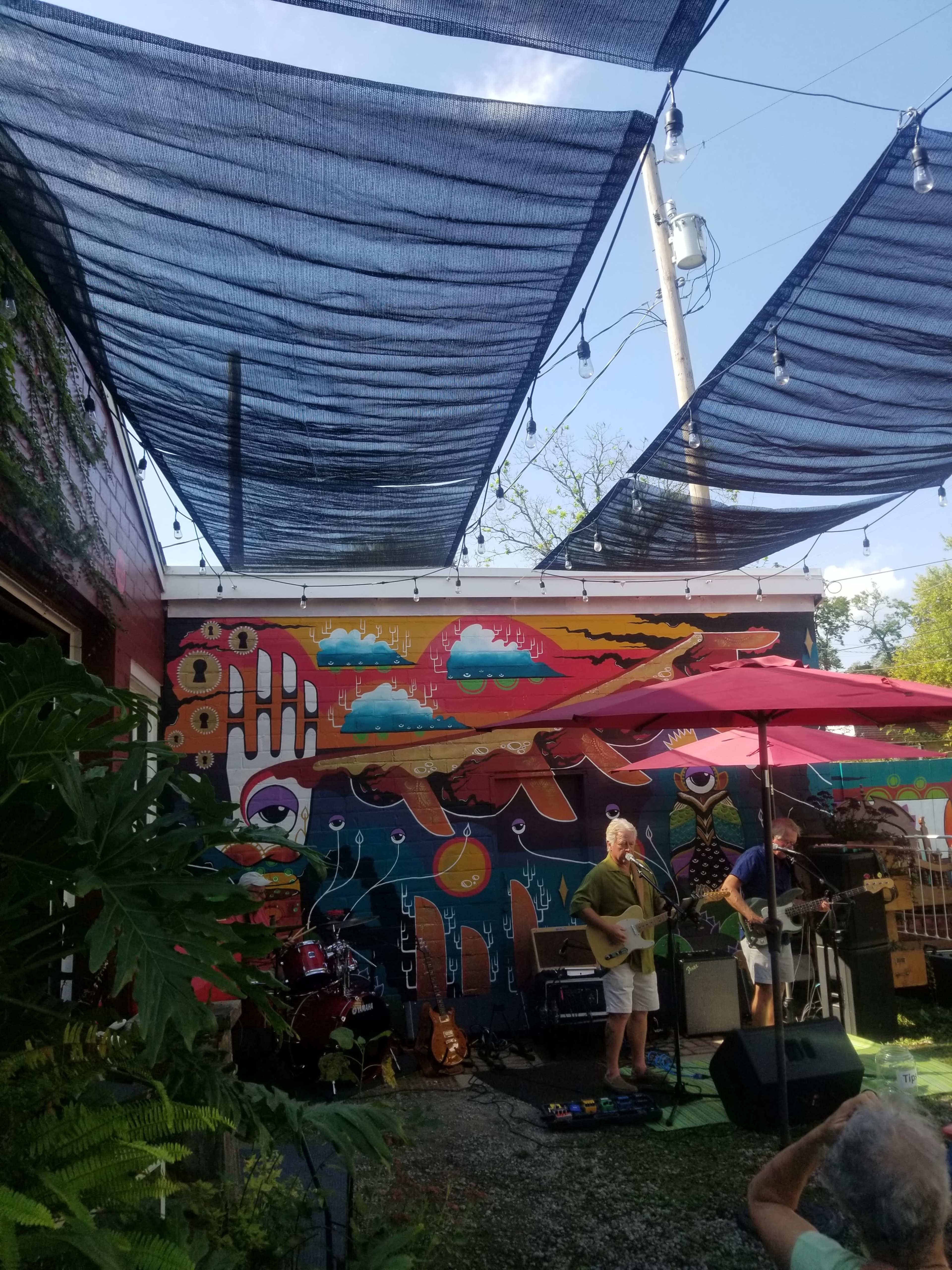 A small outdoor music performance is set up in front of a colorful mural, with shaded areas created by overhead fabric and umbrellas.