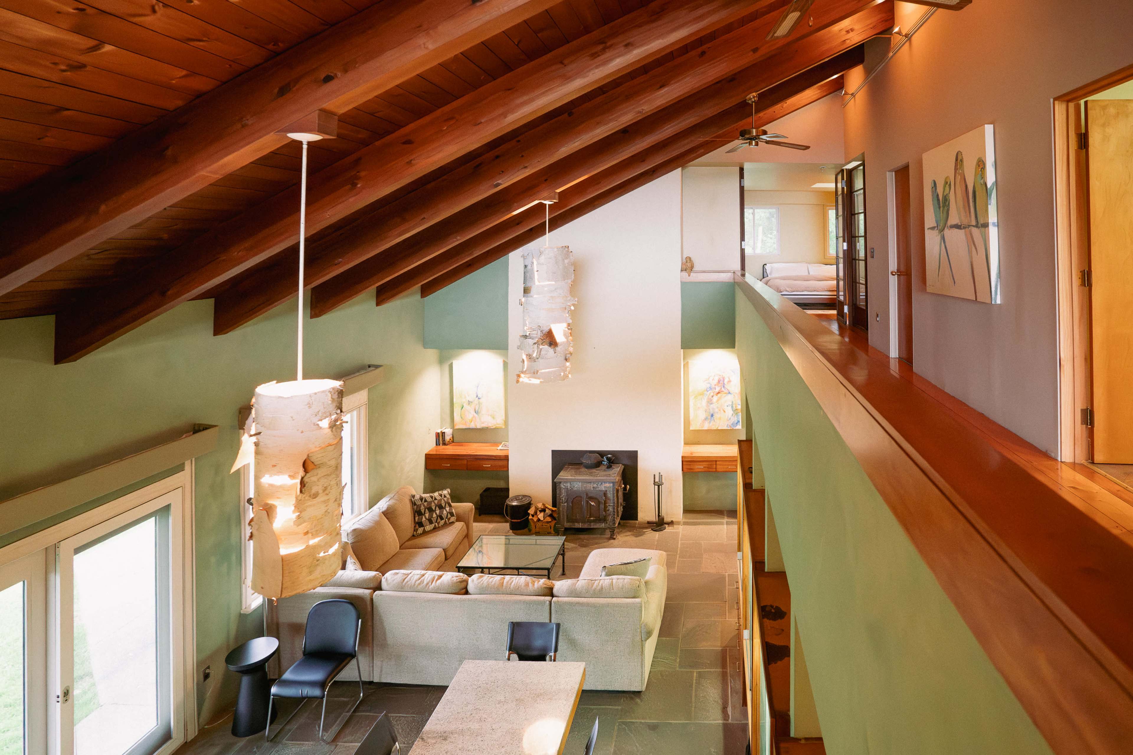 A spacious living area with a high wooden ceiling, featuring a sectional sofa, artwork on the walls, and a fireplace, all surrounded by large windows.