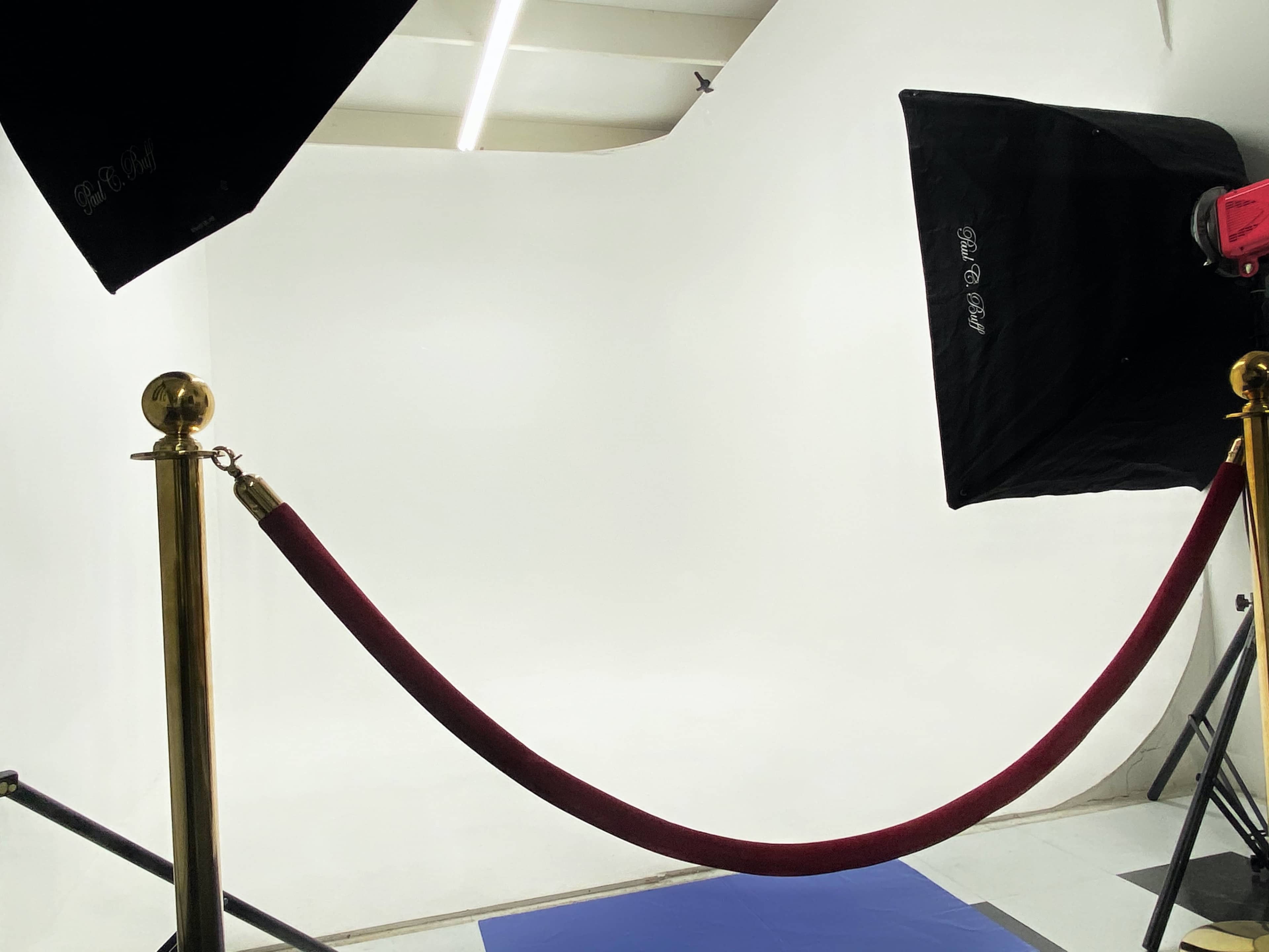 A photography setup with a white backdrop, a red velvet rope barrier, and two softbox lights positioned on either side.