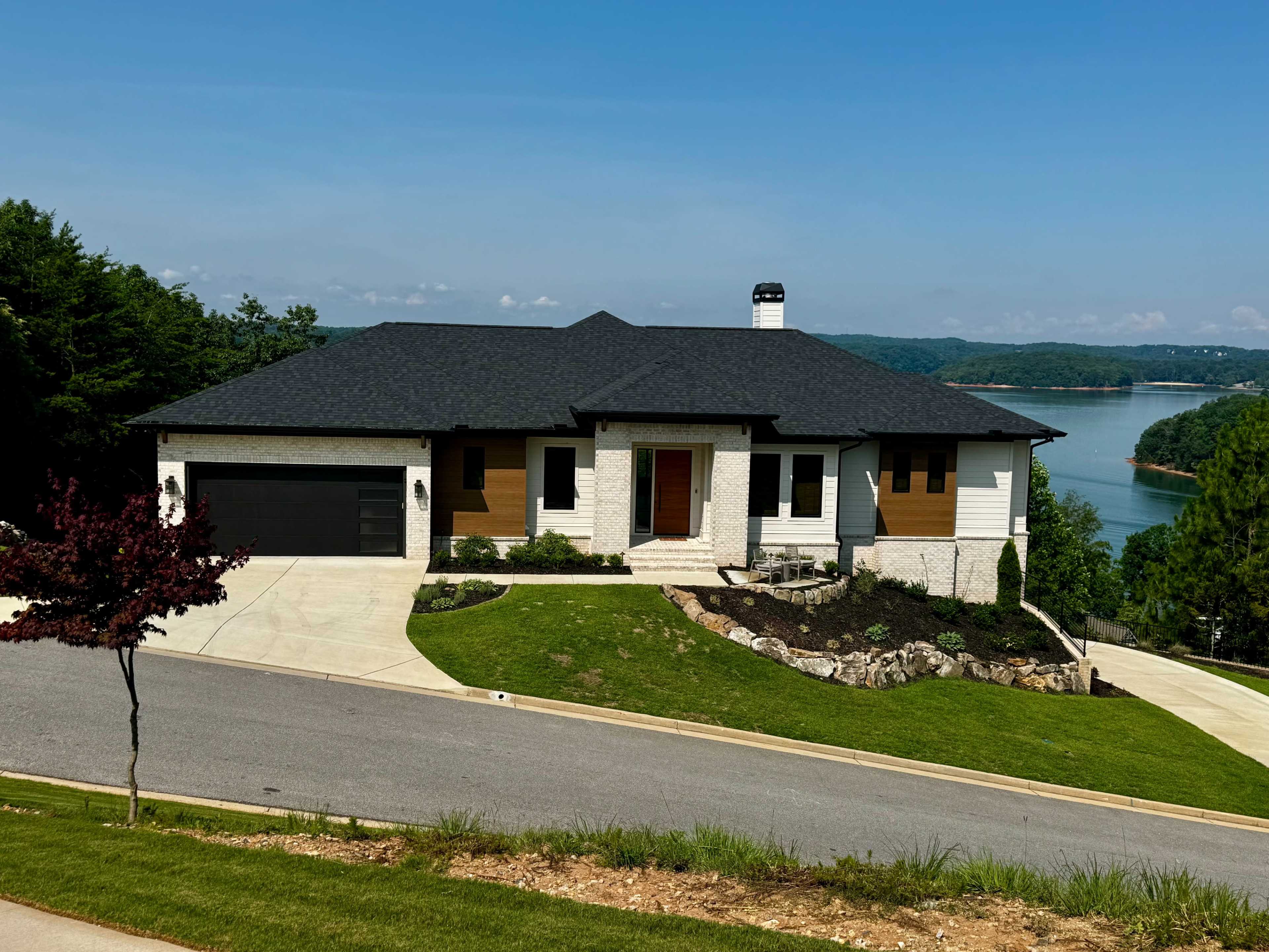 A modern single-story house with a black roof and a mix of white and wood paneling is situated on a grassy hillside overlooking a lake.
