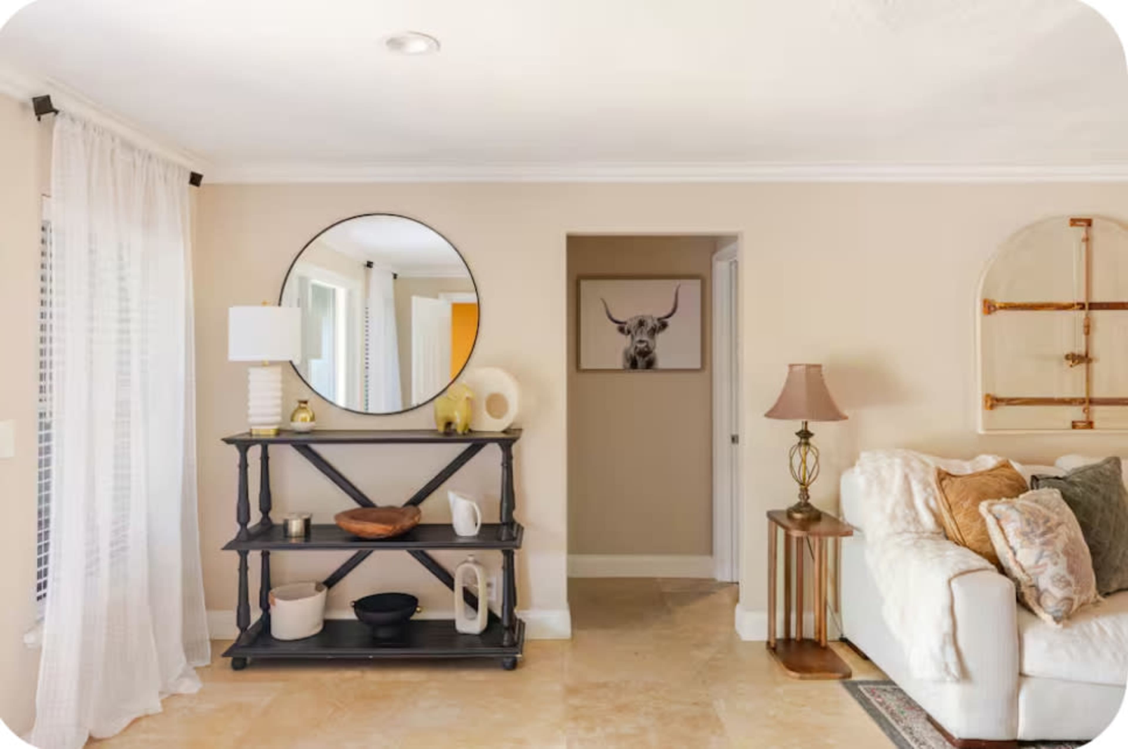 A modern living room features a circular mirror above a black shelf with various decorative items, a white sofa with cushions, and a glimpse of a doorway with artwork of a deer on the wall.