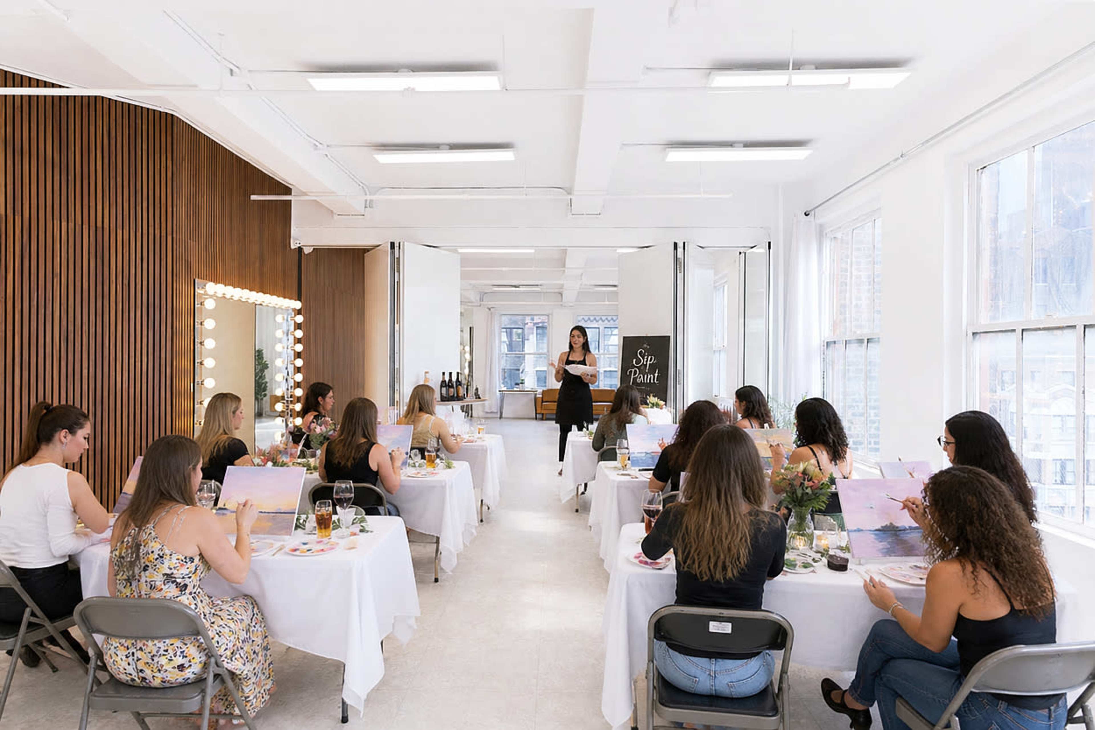 A group of people is seated at tables in a well-lit studio, engaged in a painting class led by an instructor.