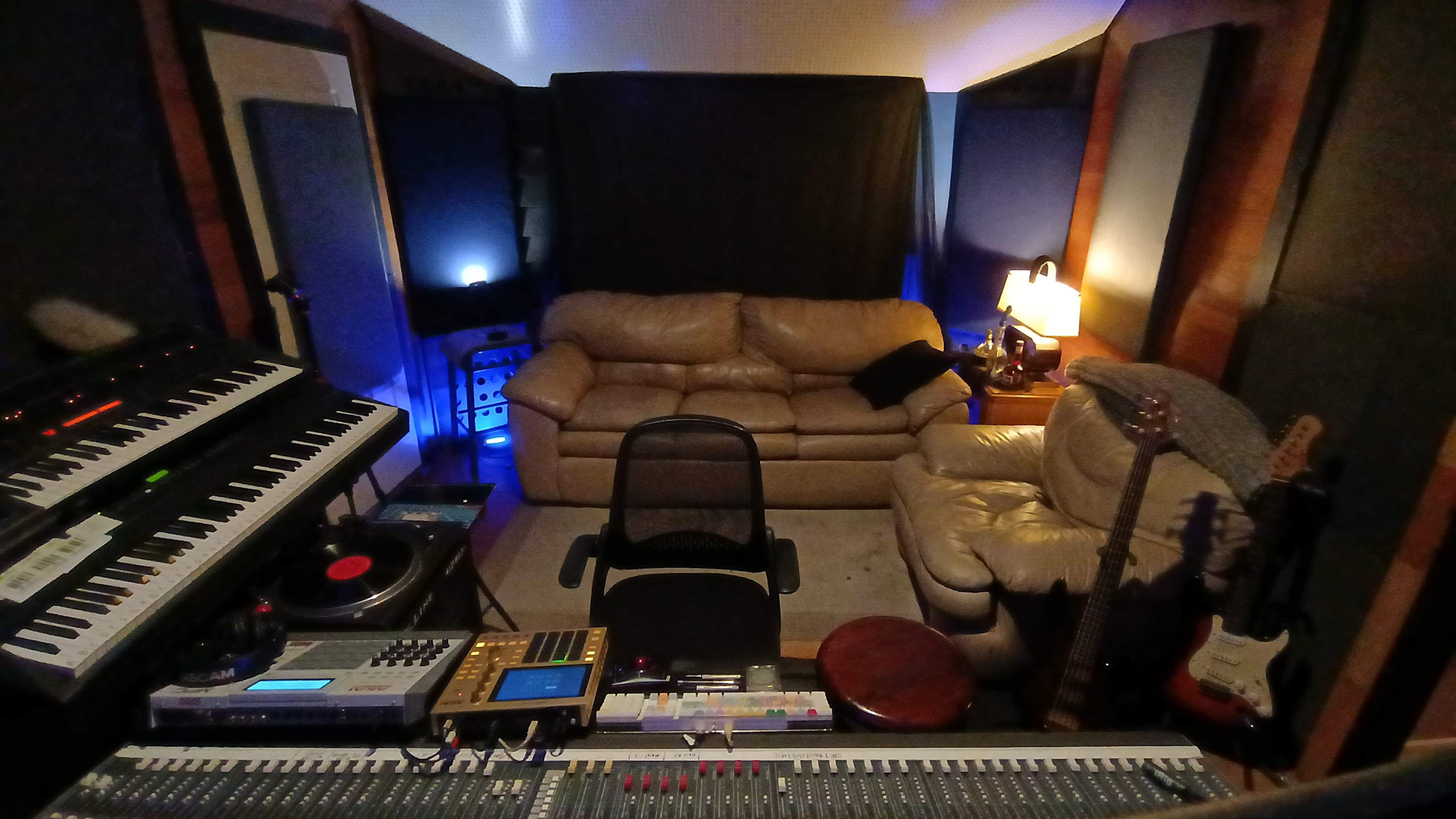 The image shows a music recording studio with a mixing console, keyboards, and two couches surrounded by soundproofing panels.