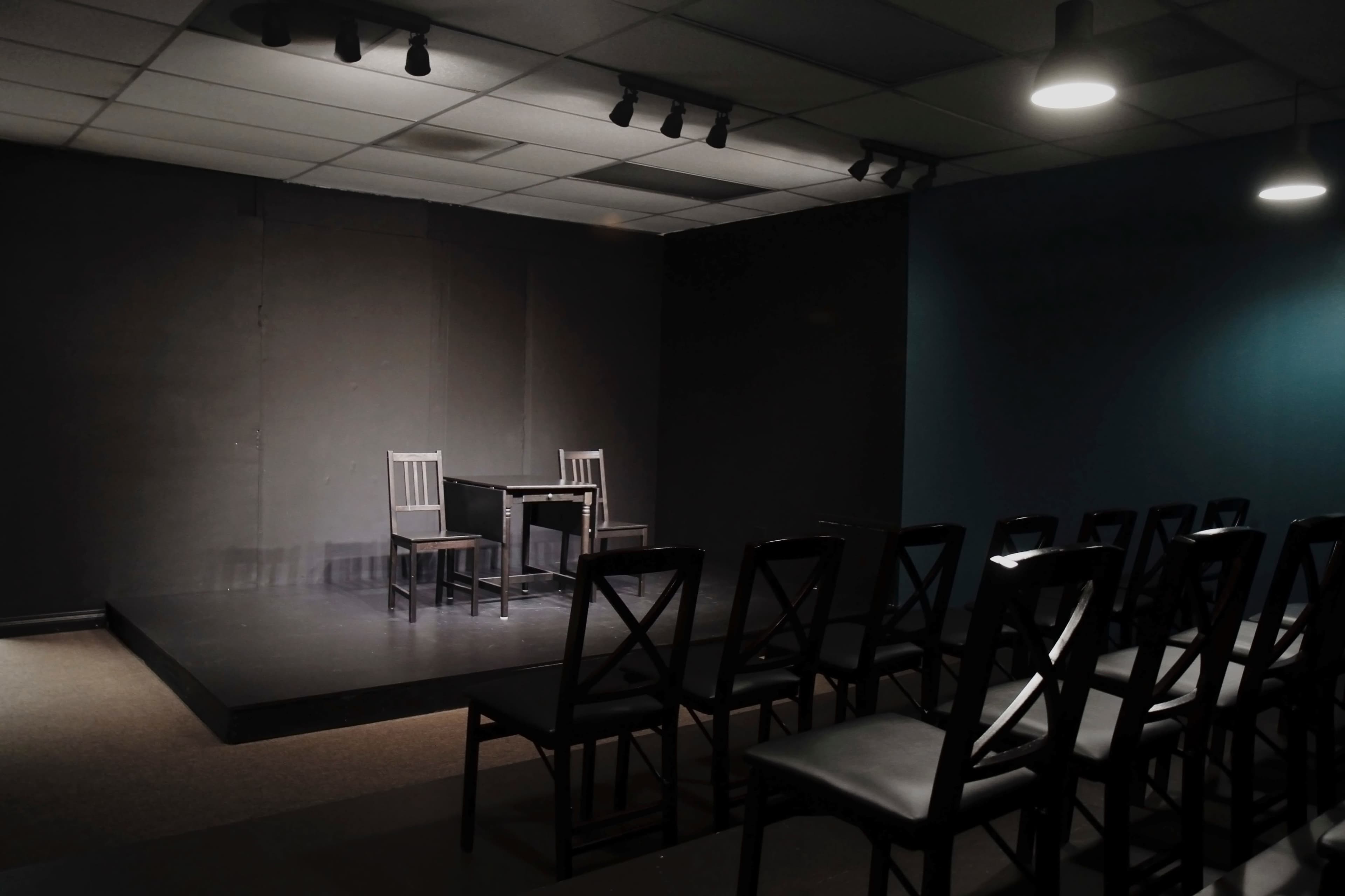 A small theater stage features two chairs and a table under dim lighting, with empty rows of black chairs facing it.