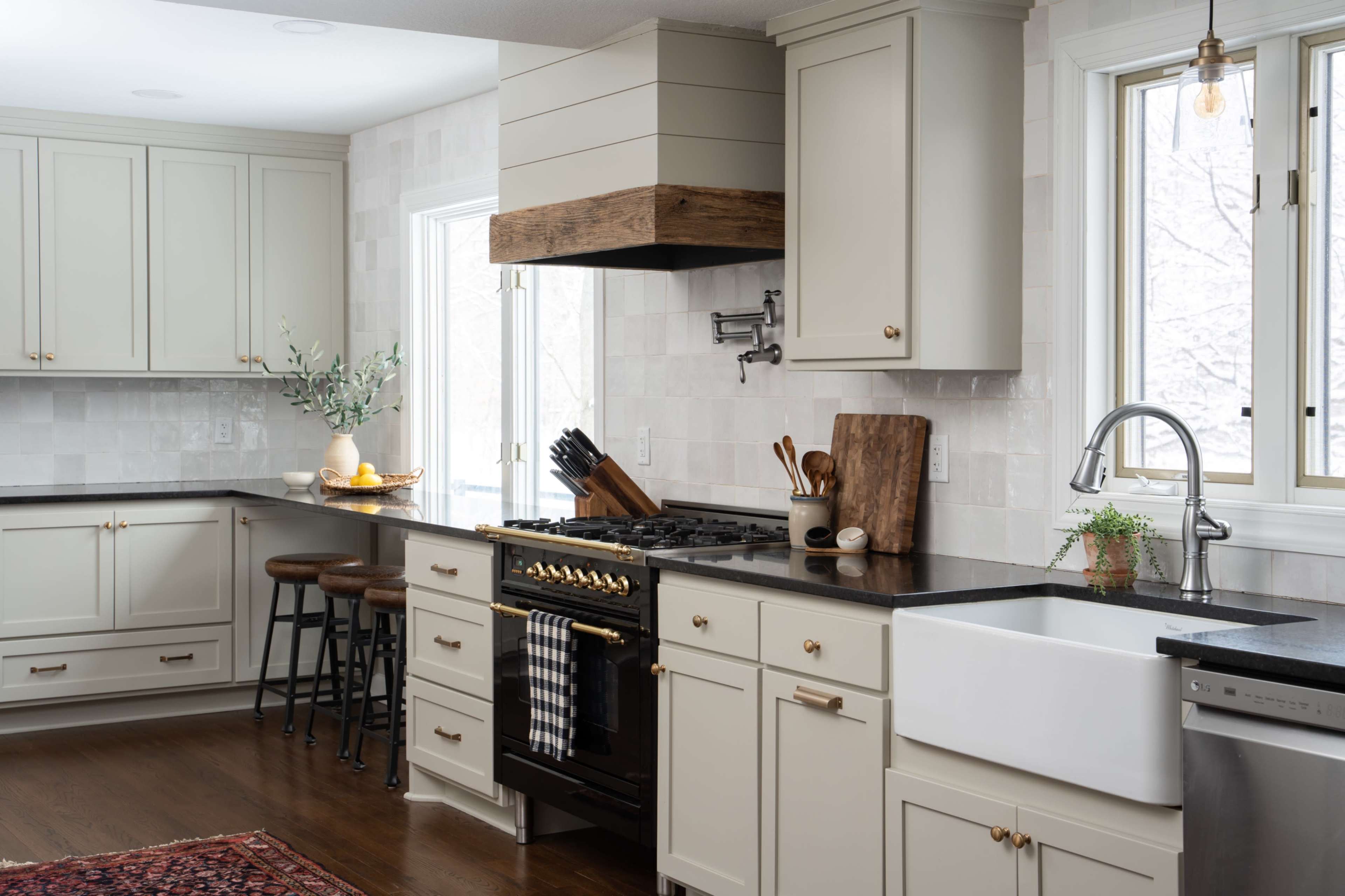 A modern kitchen features cream-colored cabinets, a large farmhouse sink, a black countertop, a gas stove with a wooden hood, and three high stools at the island.