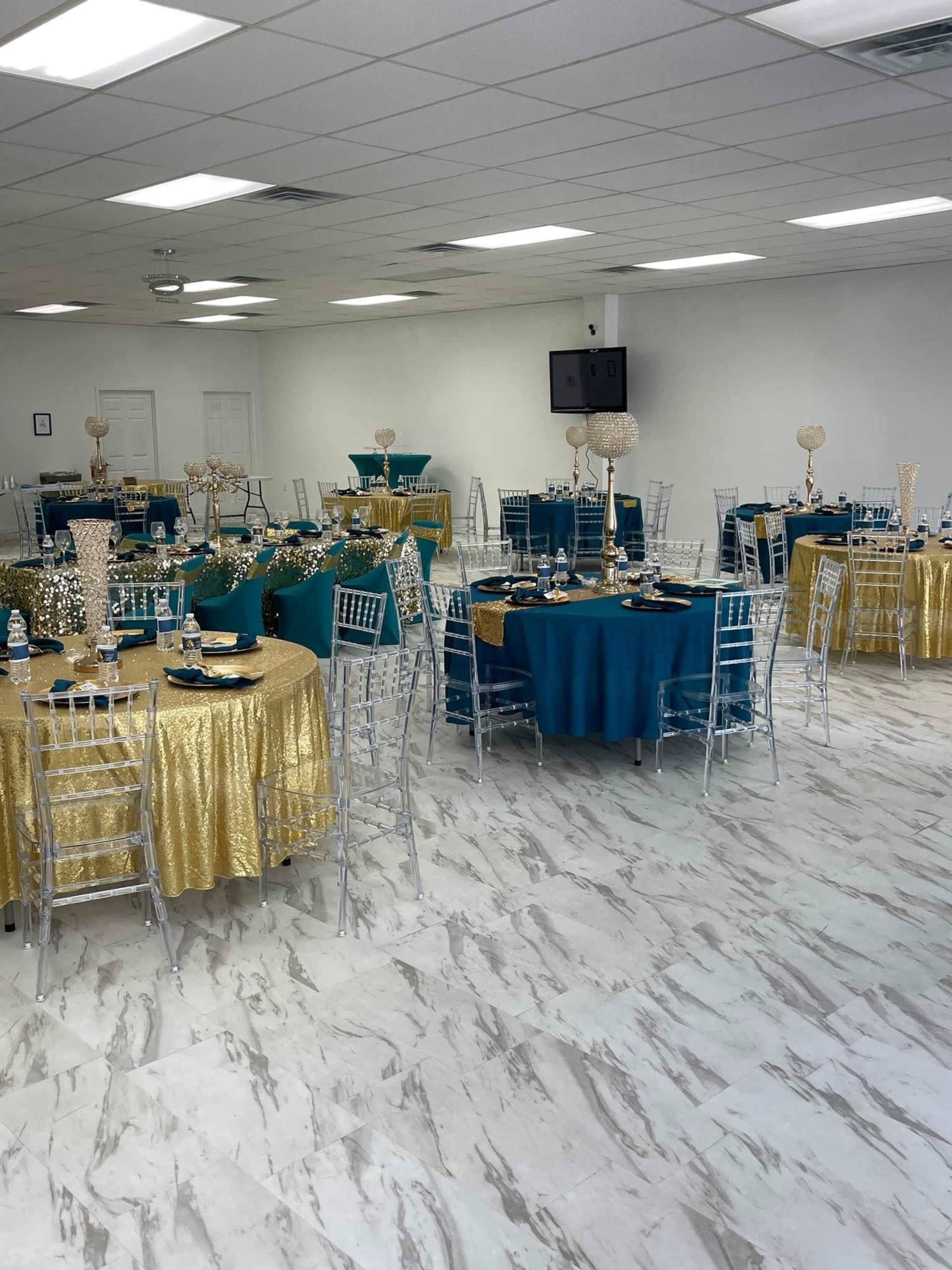 A banquet hall with several tables covered in turquoise and gold tablecloths, flanked by clear acrylic chairs.