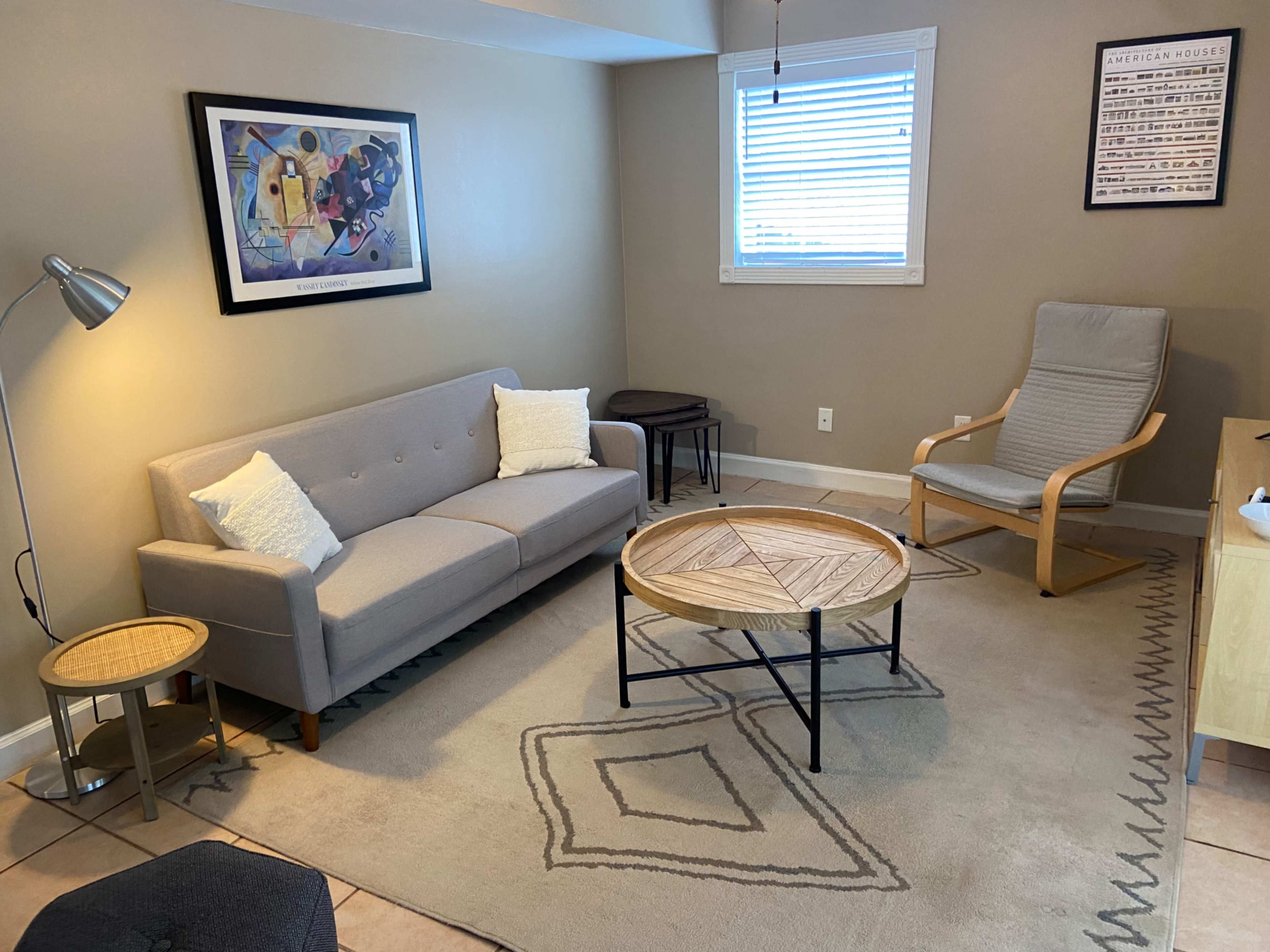 A cozy living room features a gray sofa, a wooden coffee table with a geometric design, a light-colored armchair, and a lamp, set against a neutral wall.