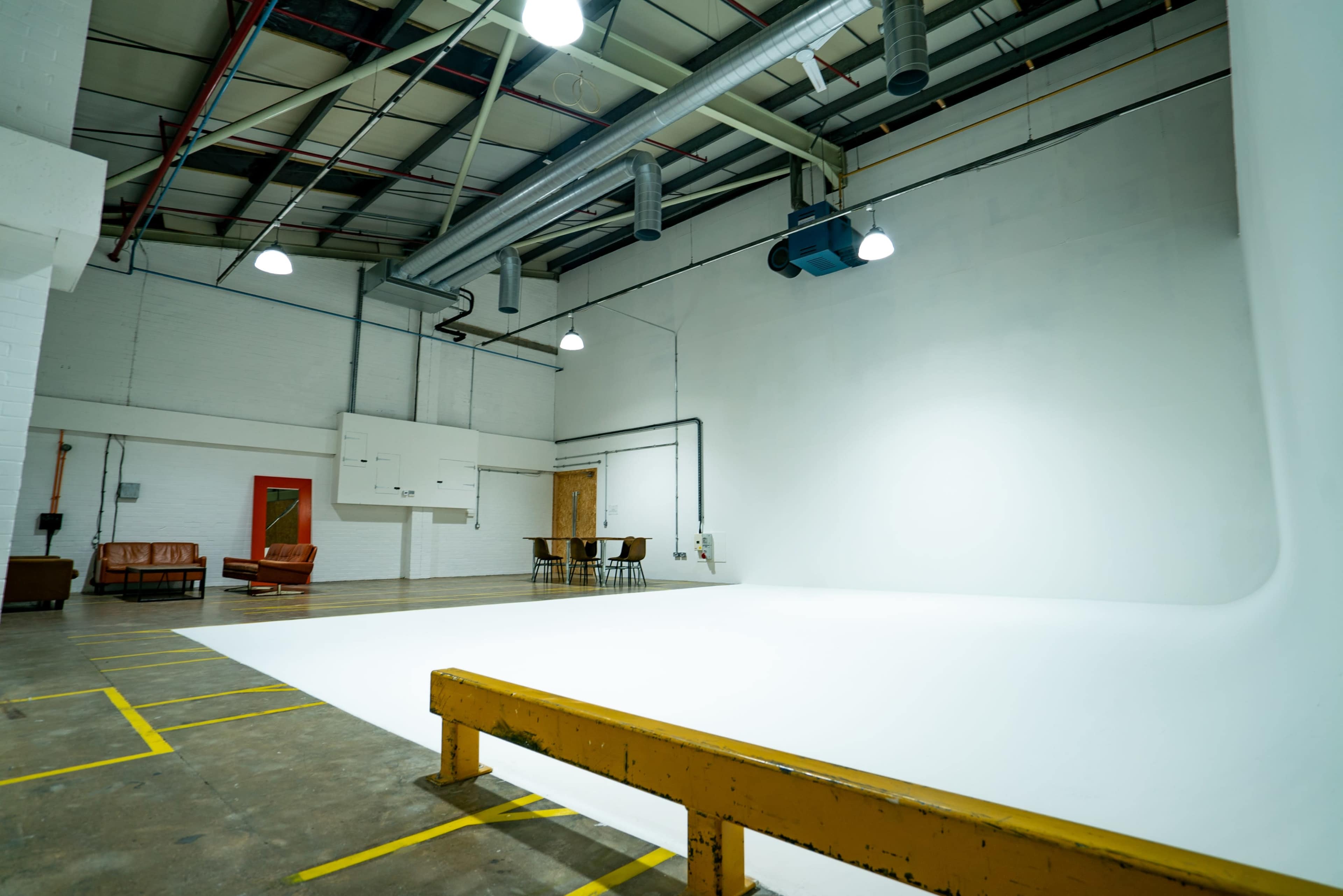 The image shows an empty studio space with a white backdrop, a wooden bench, a table with chairs, and overhead lights.