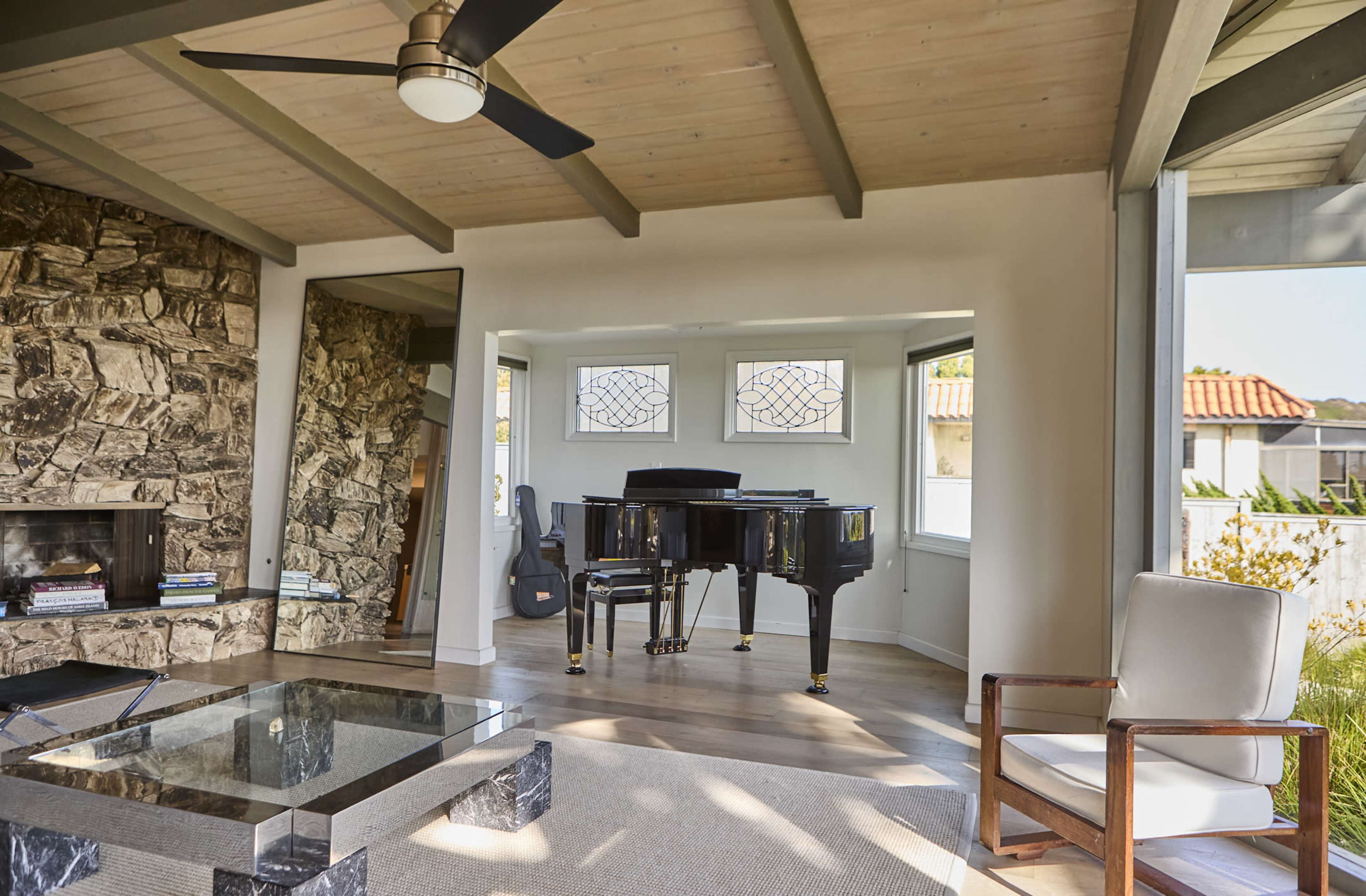 A black grand piano is positioned in a well-lit room with large windows and a stone wall.