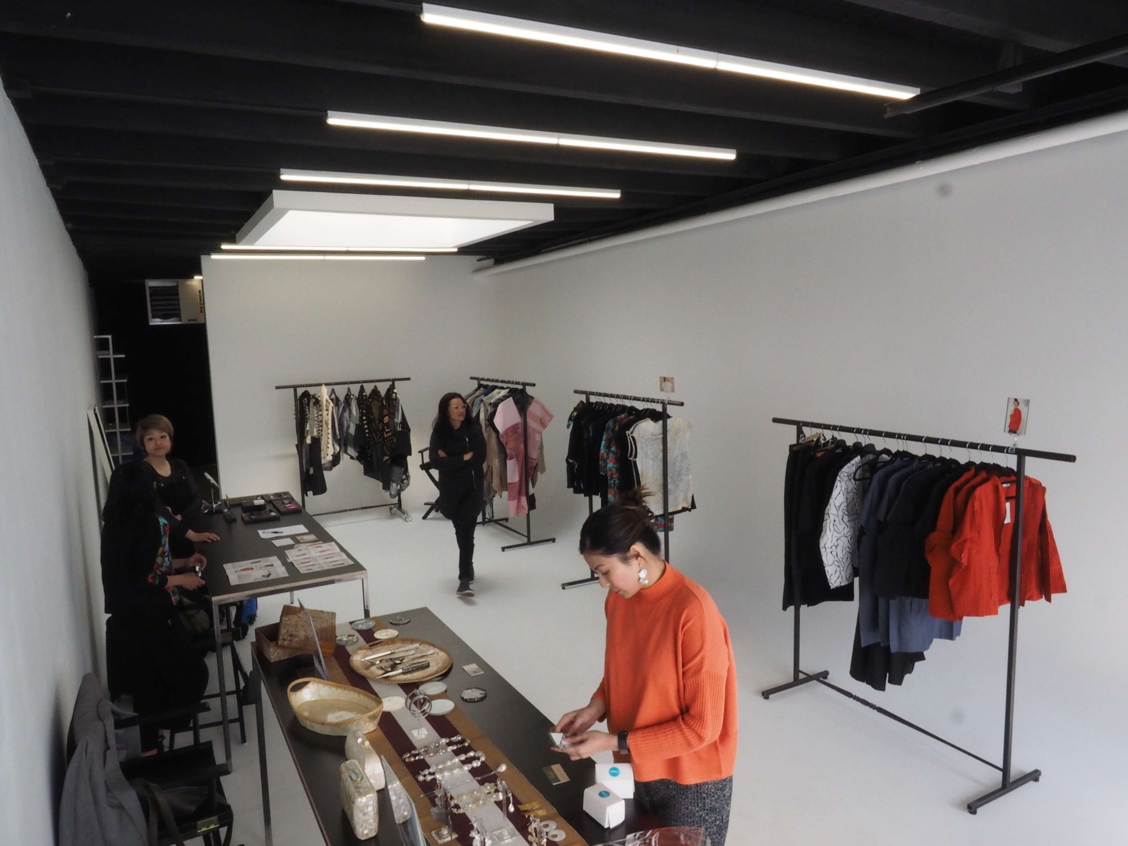 A jewelry and clothing boutique features a display table with accessories and multiple clothing racks against a minimalist white backdrop.