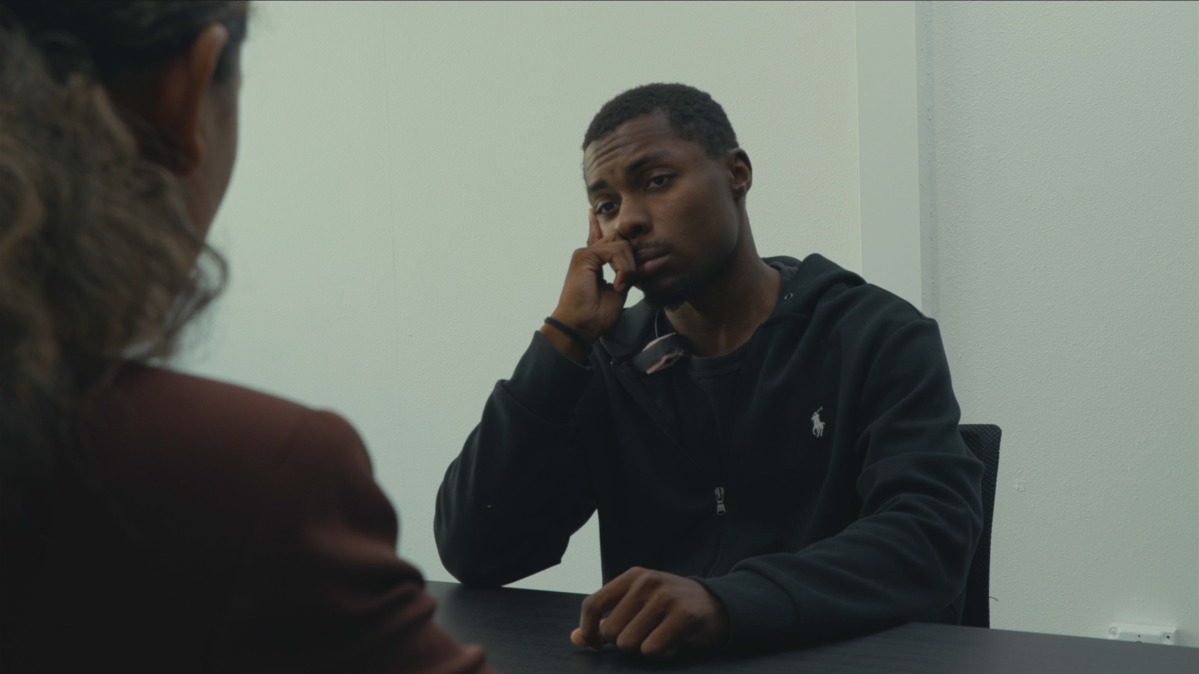 A young man sits at a table, resting his chin on his hand, while looking towards a person off-screen.