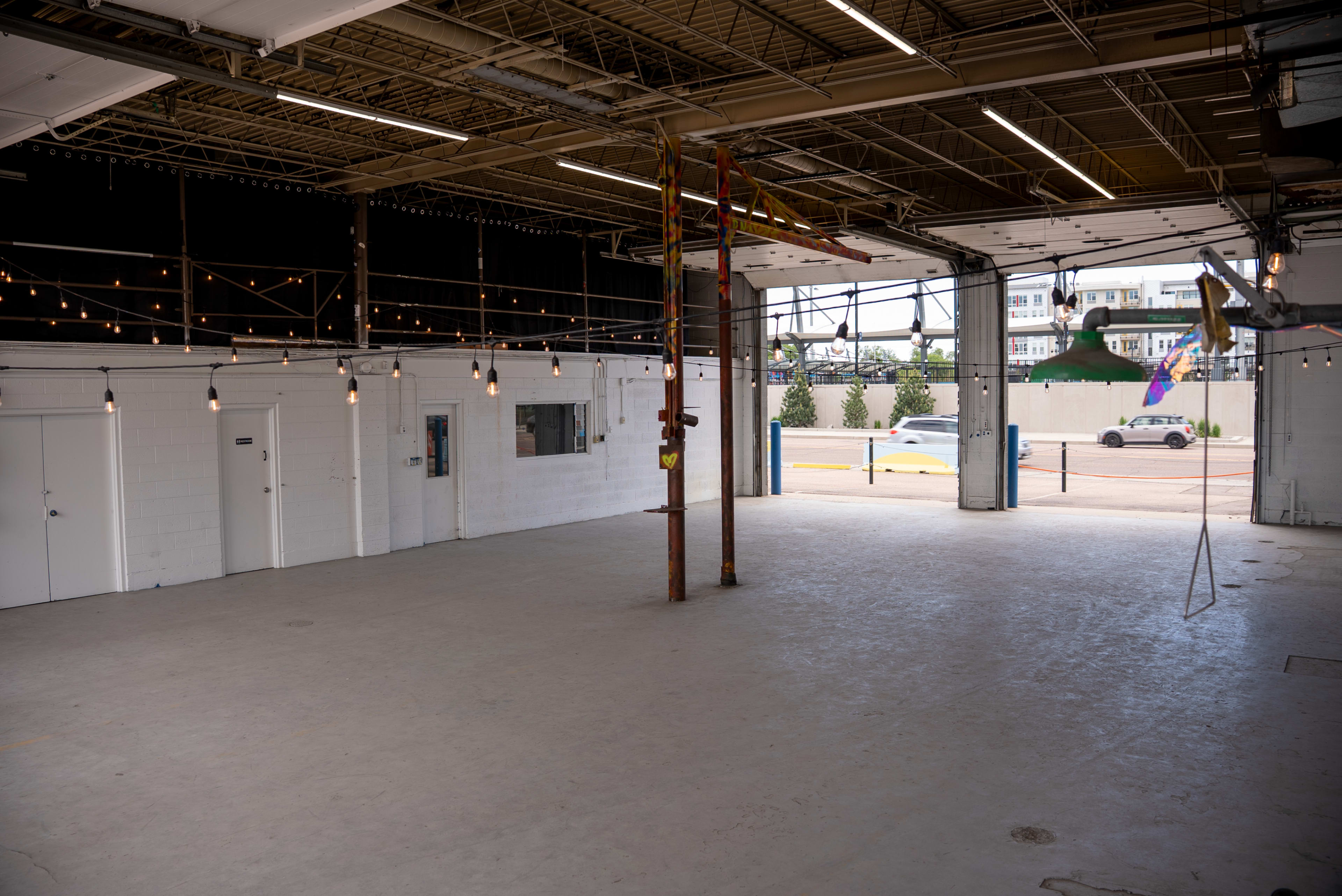 The image shows an empty industrial space with concrete flooring, string lights, and an open garage door leading to a street outside.