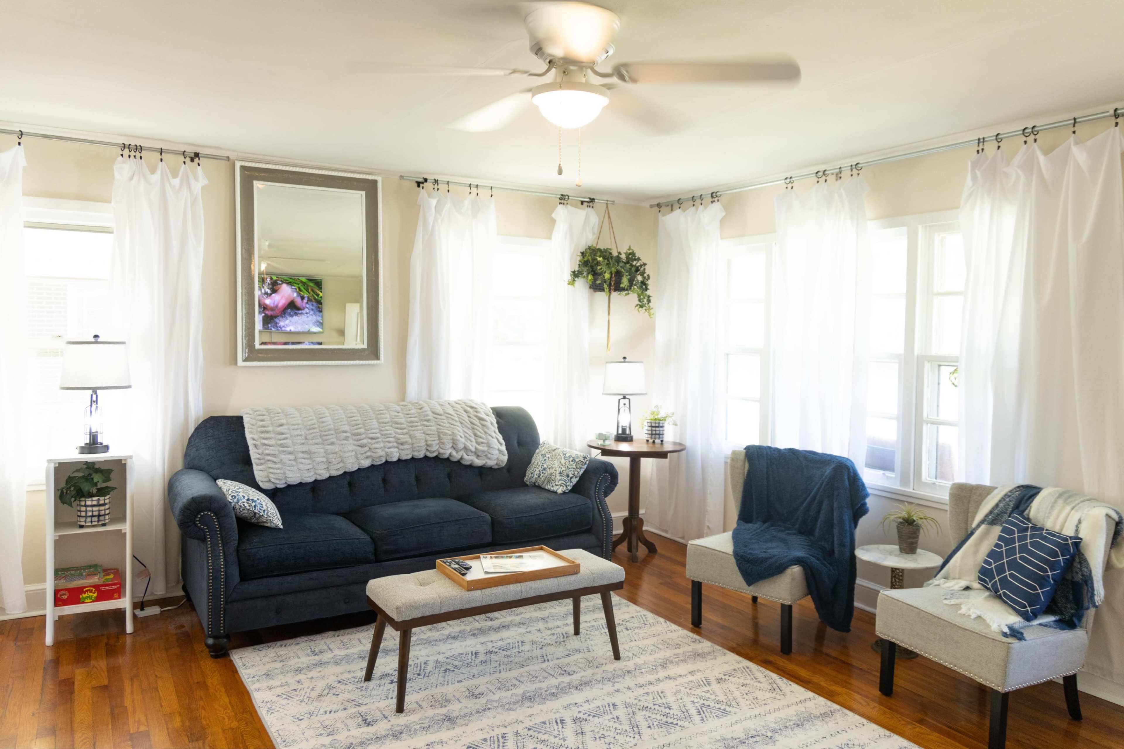 A bright living room features a blue sofa, two armchairs, a coffee table, and large windows adorned with white curtains.