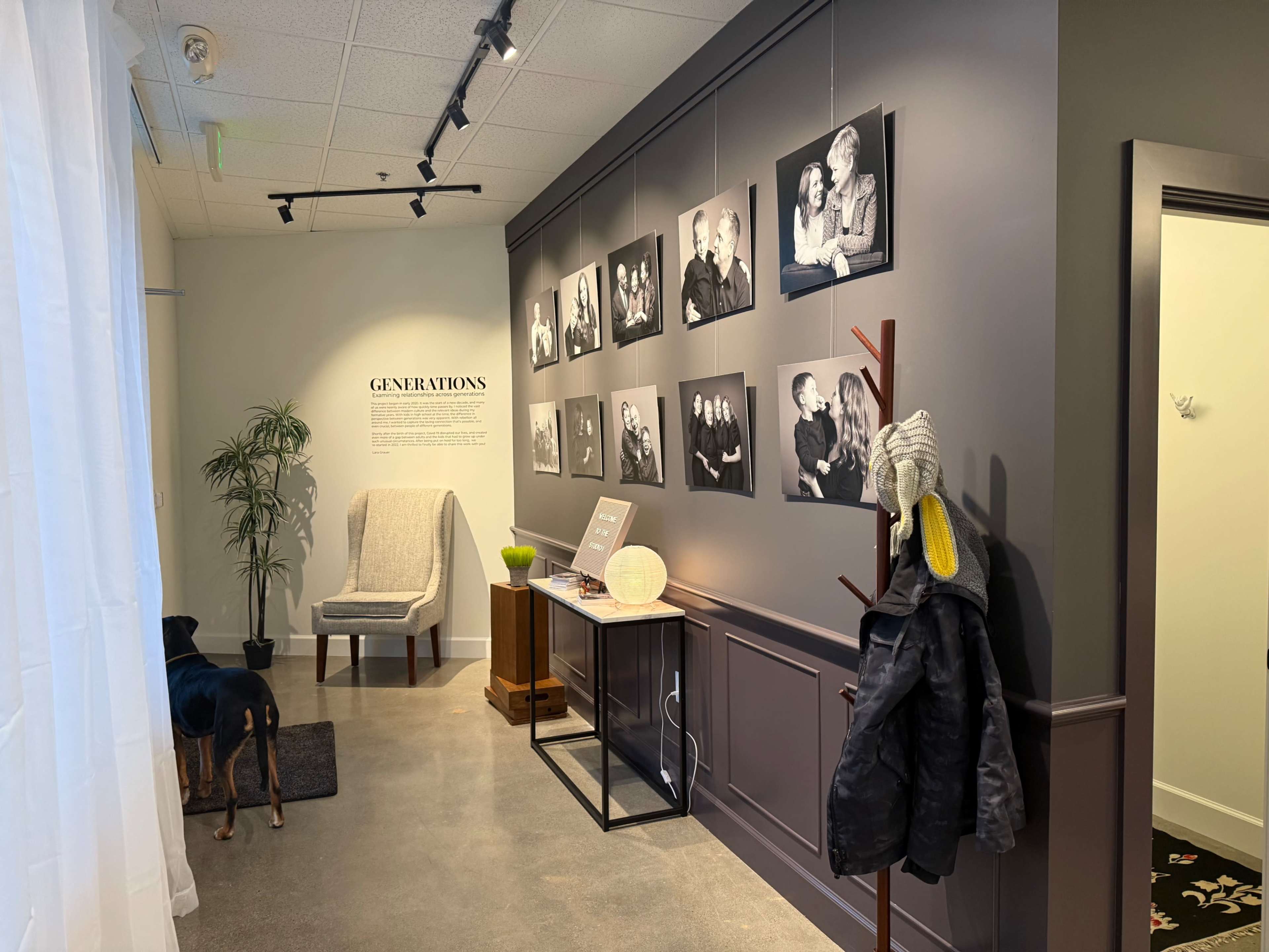 The image shows a gallery space with a wall displaying black-and-white photographs of people, alongside a chair, a table with objects, a plant, and a coat rack.