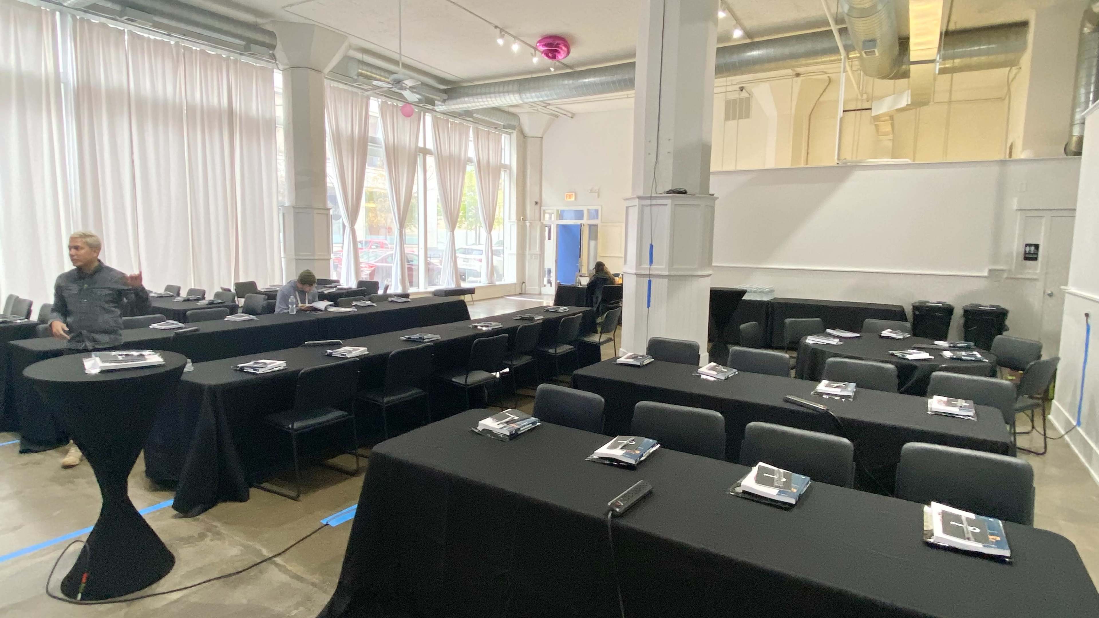 A spacious room set up for a meeting, featuring multiple rows of black-covered tables with chairs, and large windows with sheer curtains.