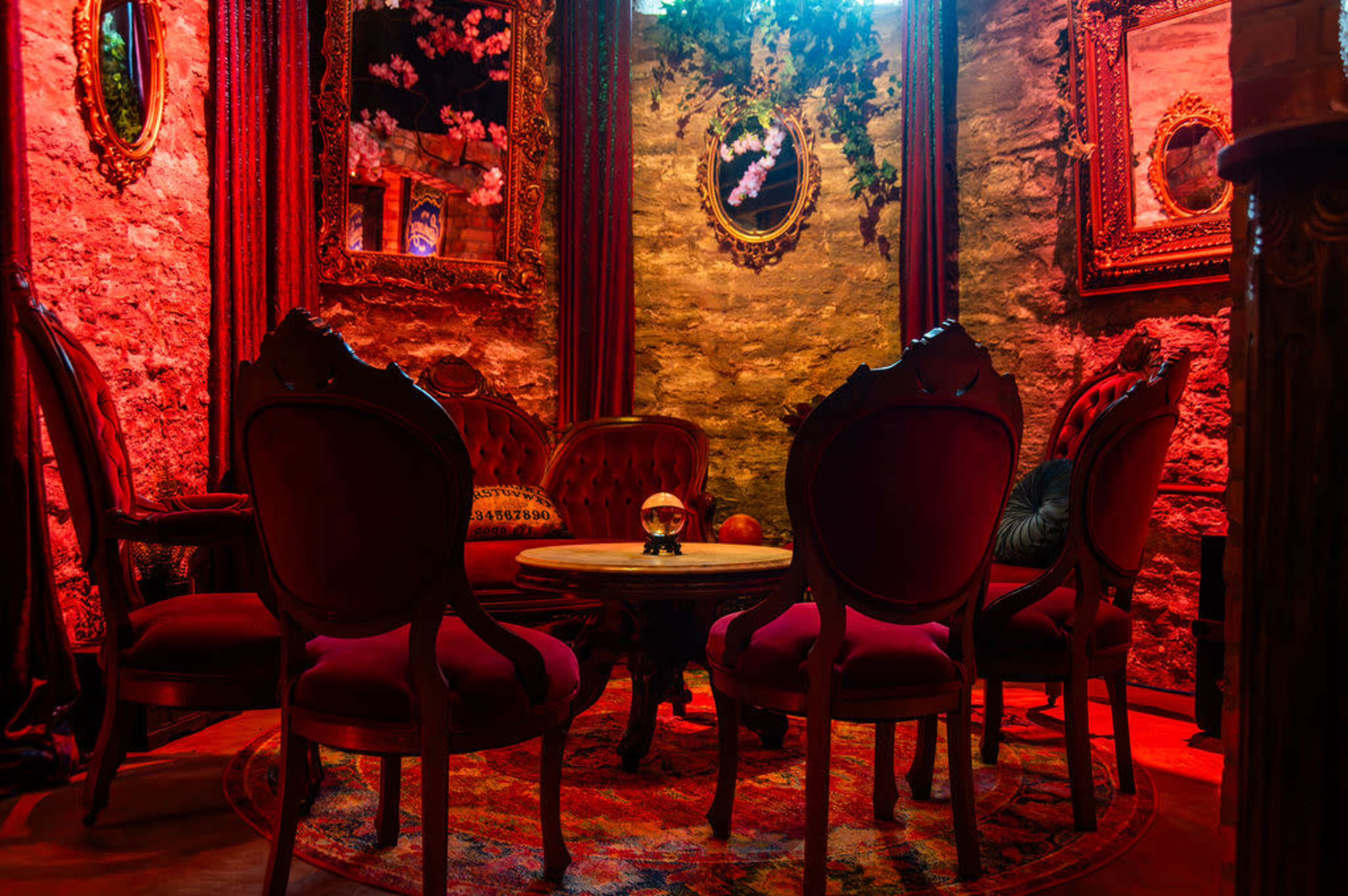 A dimly lit room features ornate red chairs arranged around a wooden table, with stone walls and decorative mirrors.