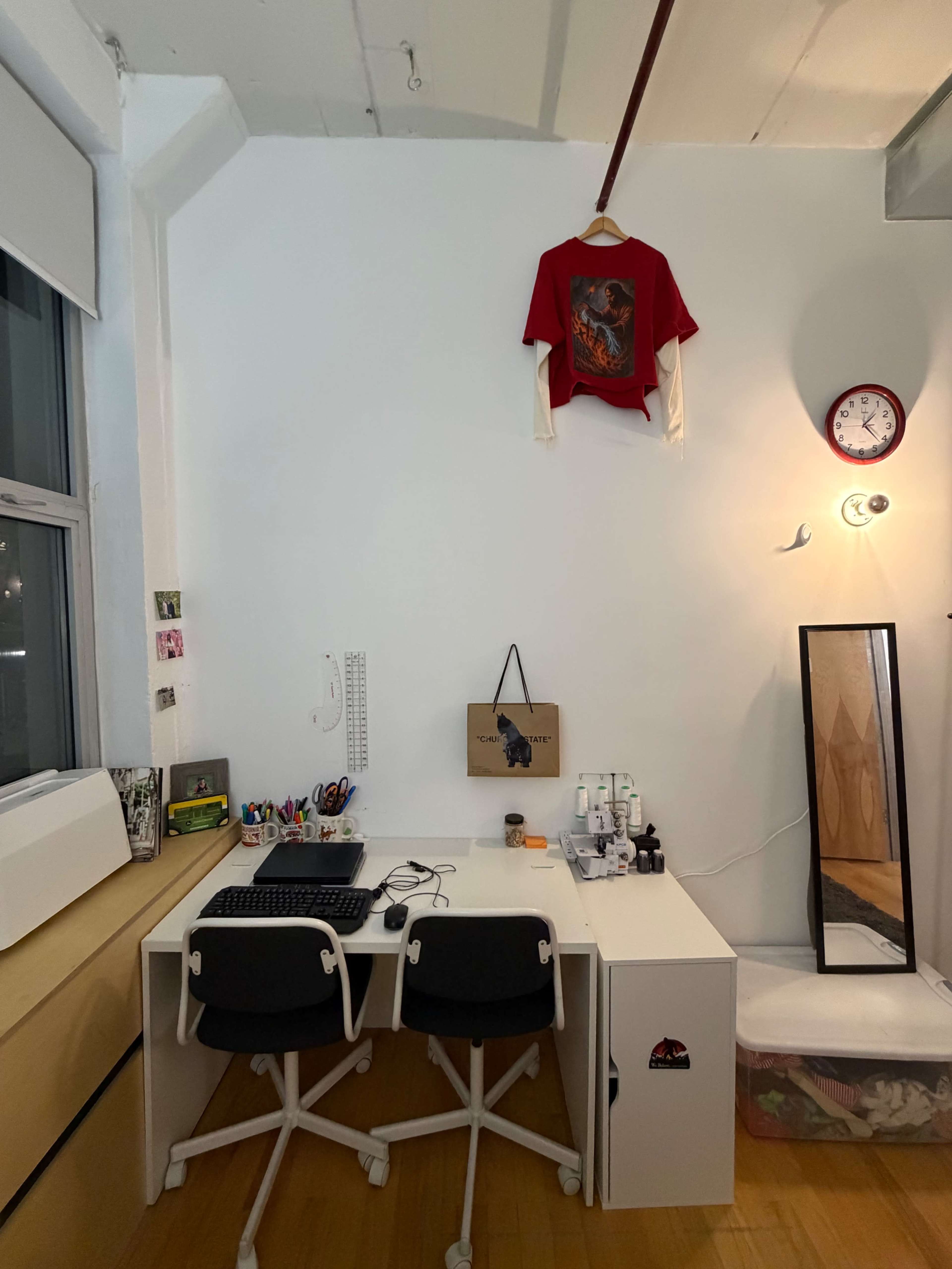 A small workspace features a white desk with a computer, two chairs, a clock on the wall, and a shirt hanging above it.