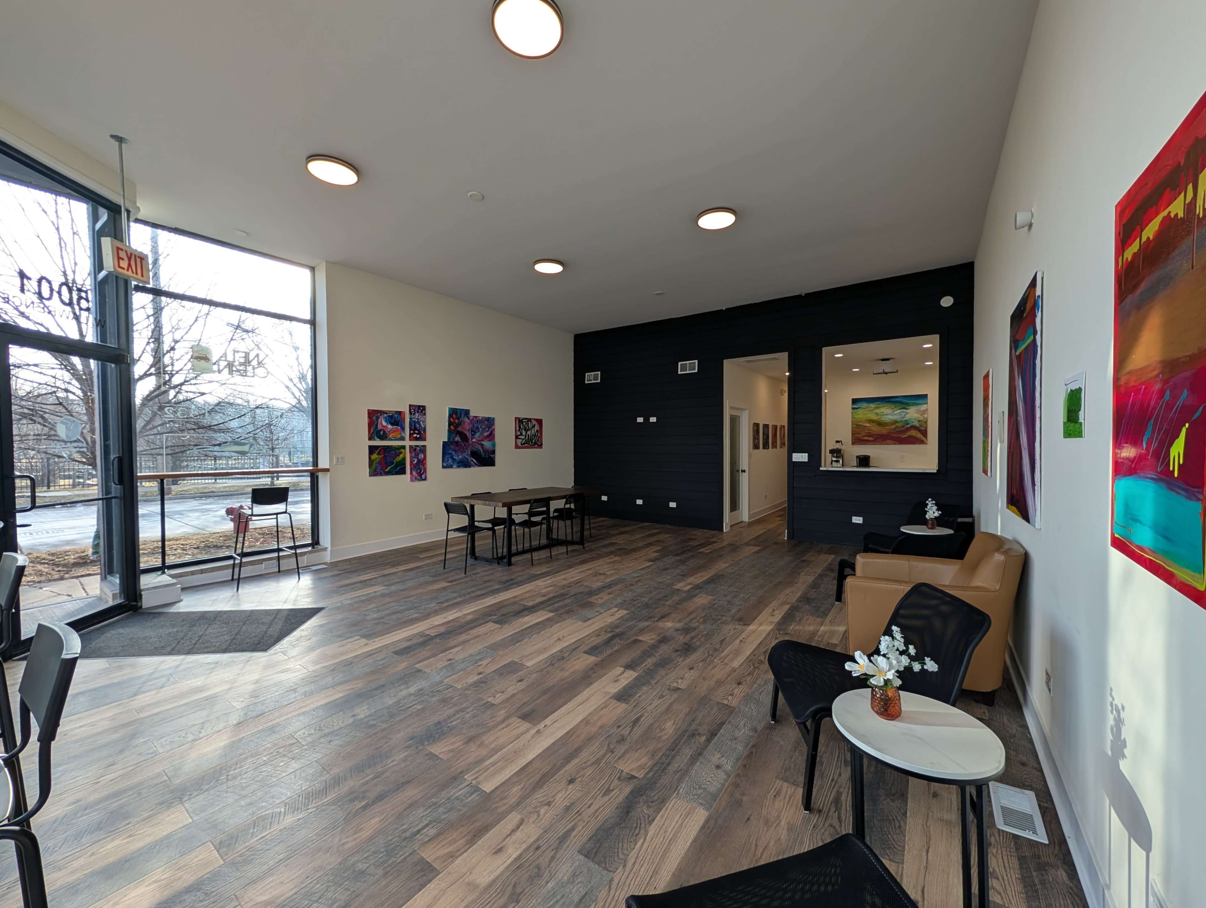 The image shows a modern, open space with wooden floors, art on the walls, and a few chairs and tables arranged for seating.