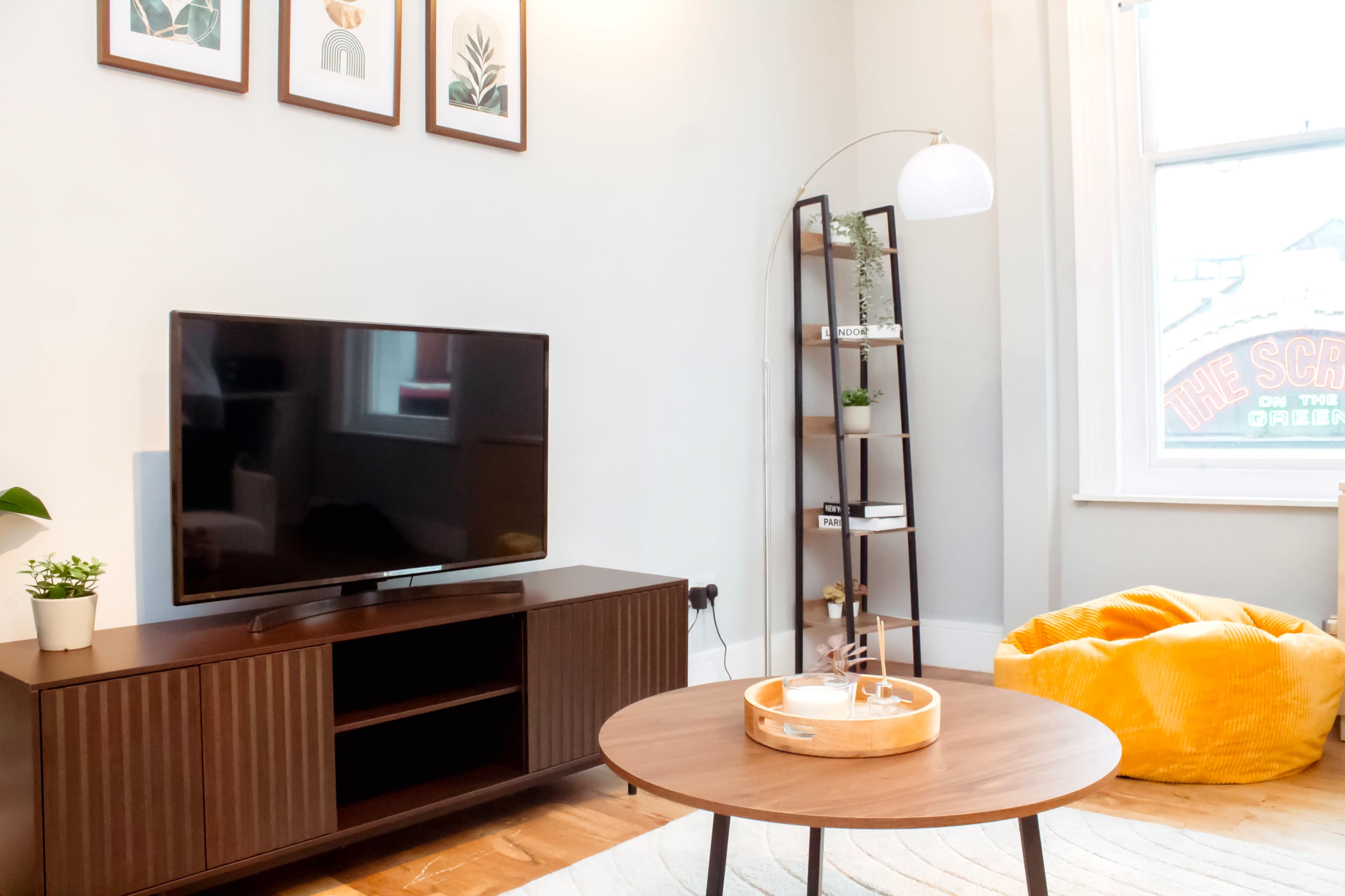 The image shows a modern living room featuring a television on a wooden cabinet, a round coffee table, a yellow bean bag, and a tall bookshelf with decorative items.