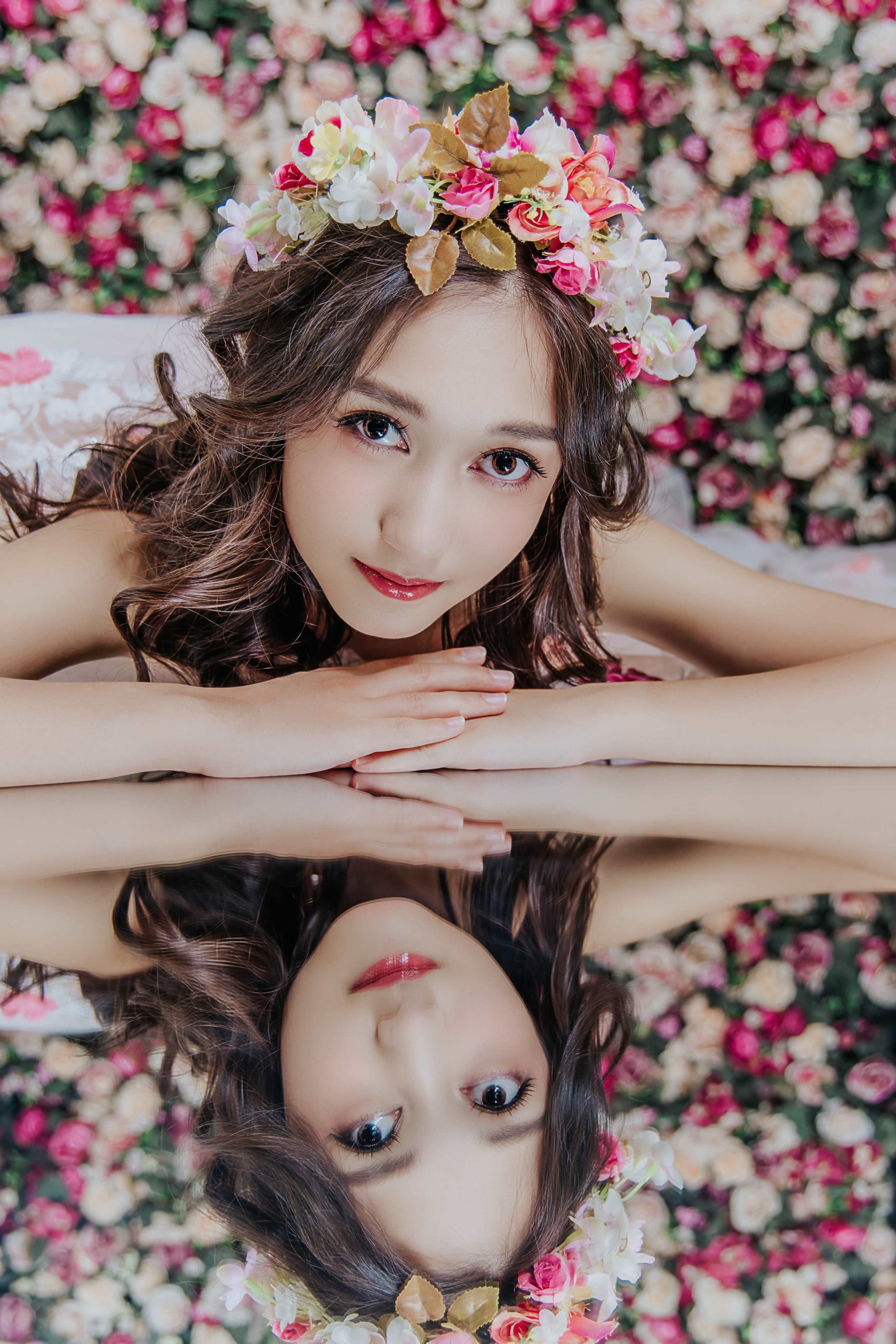 A woman with long, wavy hair and a floral crown rests her arms on a reflective surface, surrounded by a backdrop of pink and cream roses.