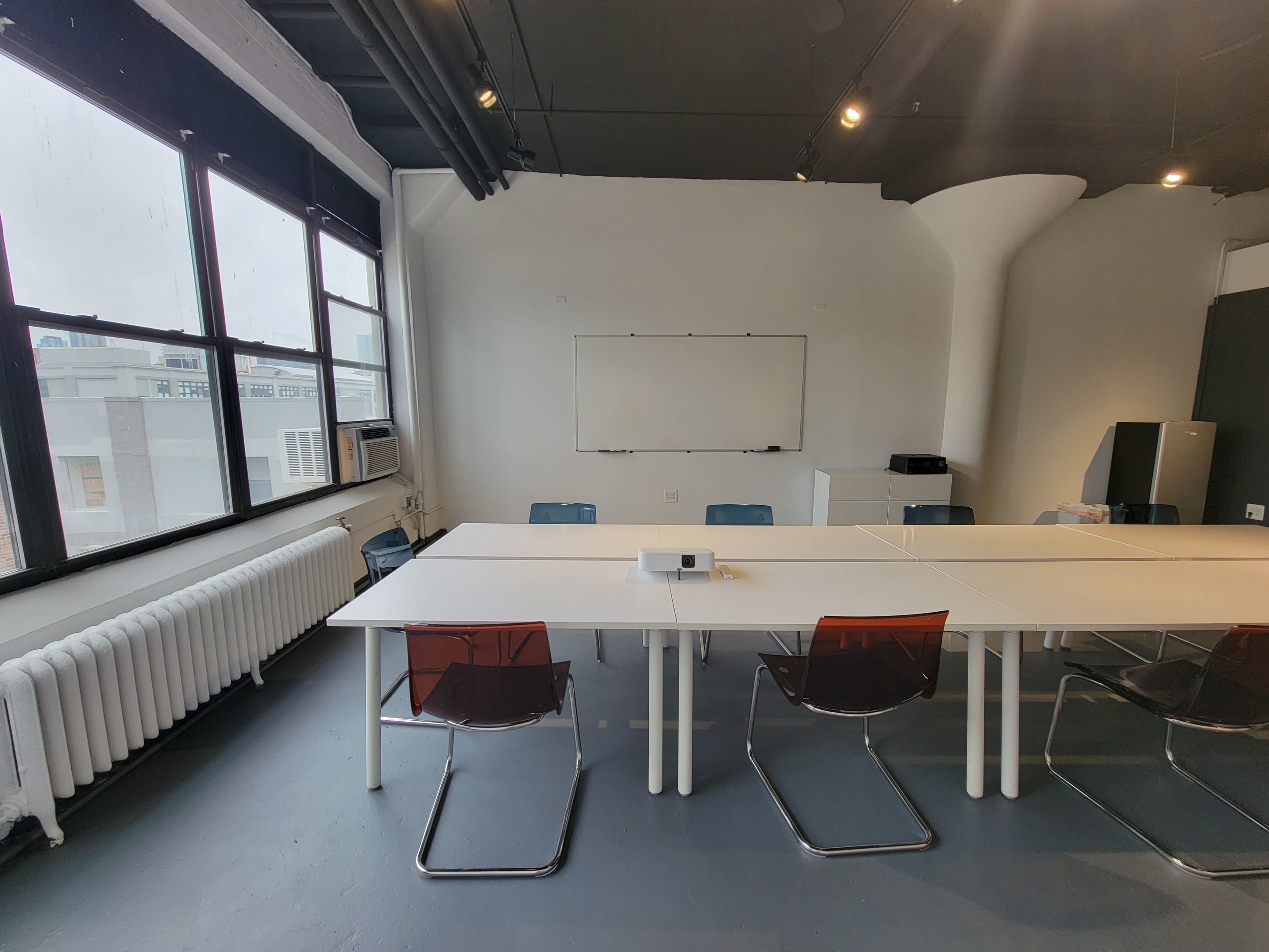 A spacious meeting room features a long table with several chairs, large windows providing natural light, and a whiteboard on the wall.