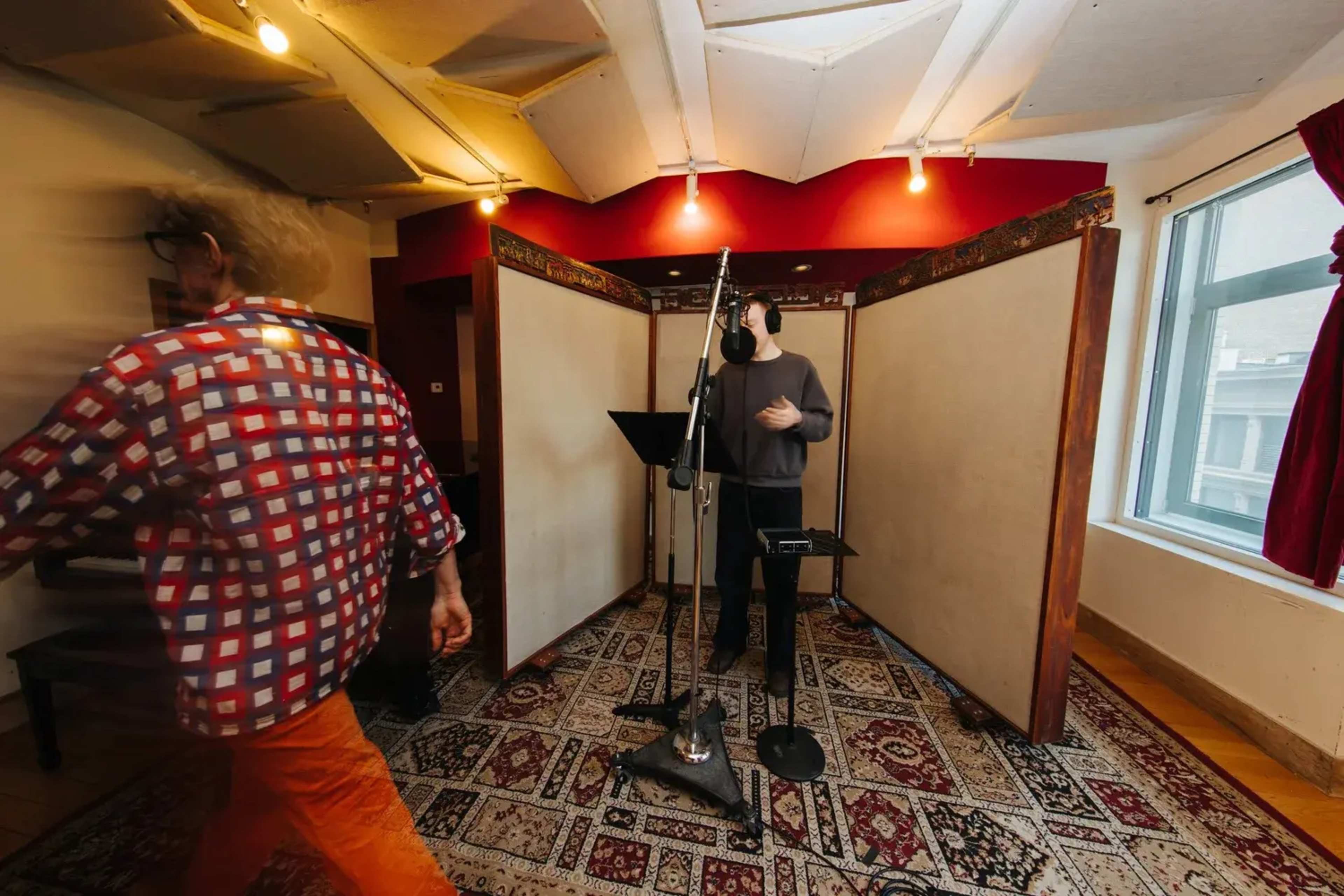A person wearing orange pants walks past a microphone setup in a sound recording studio with acoustic panels and warm lighting.