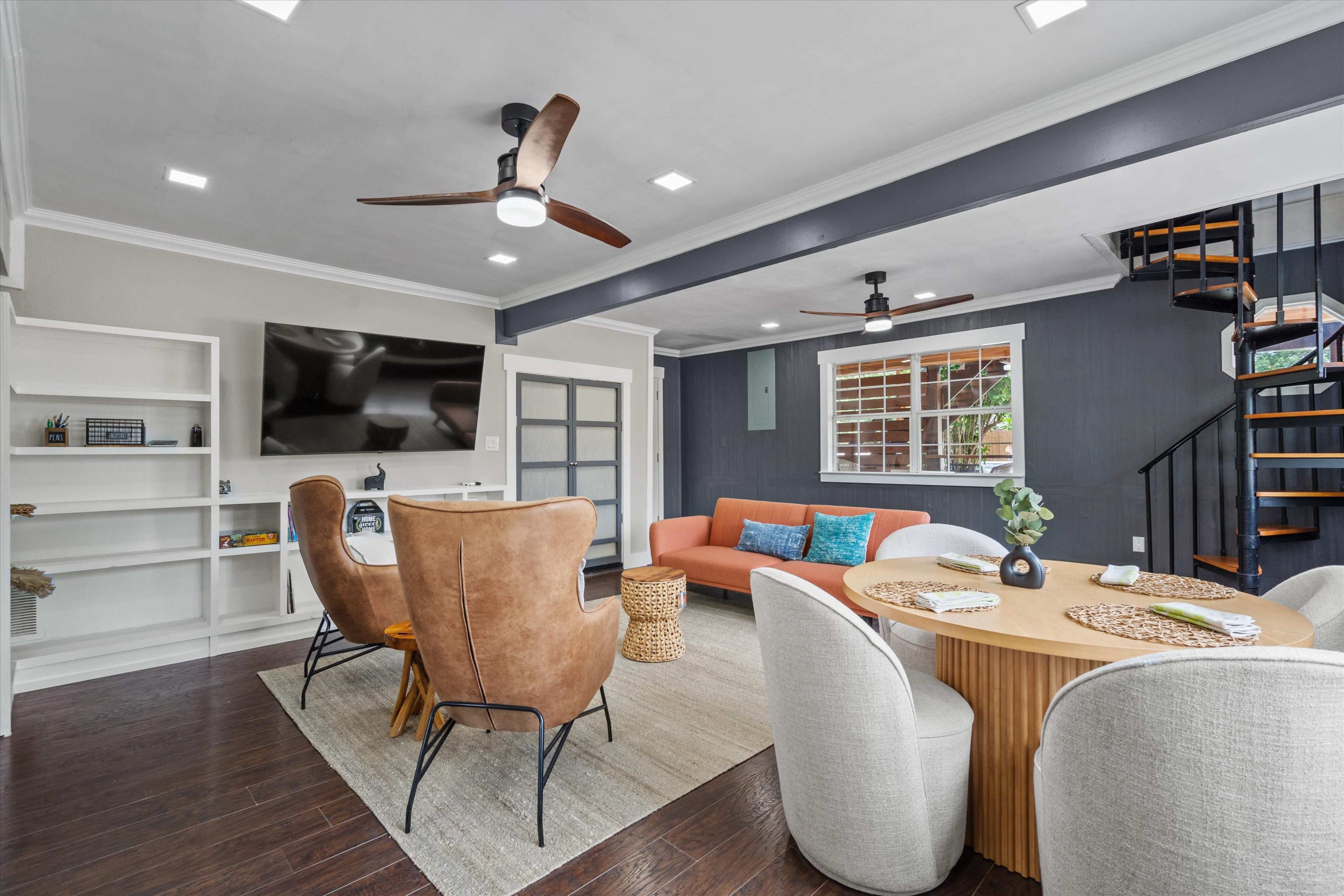 The image shows a modern living space with a mix of seating options, a round dining table, a large television mounted on the wall, and a staircase in the background.