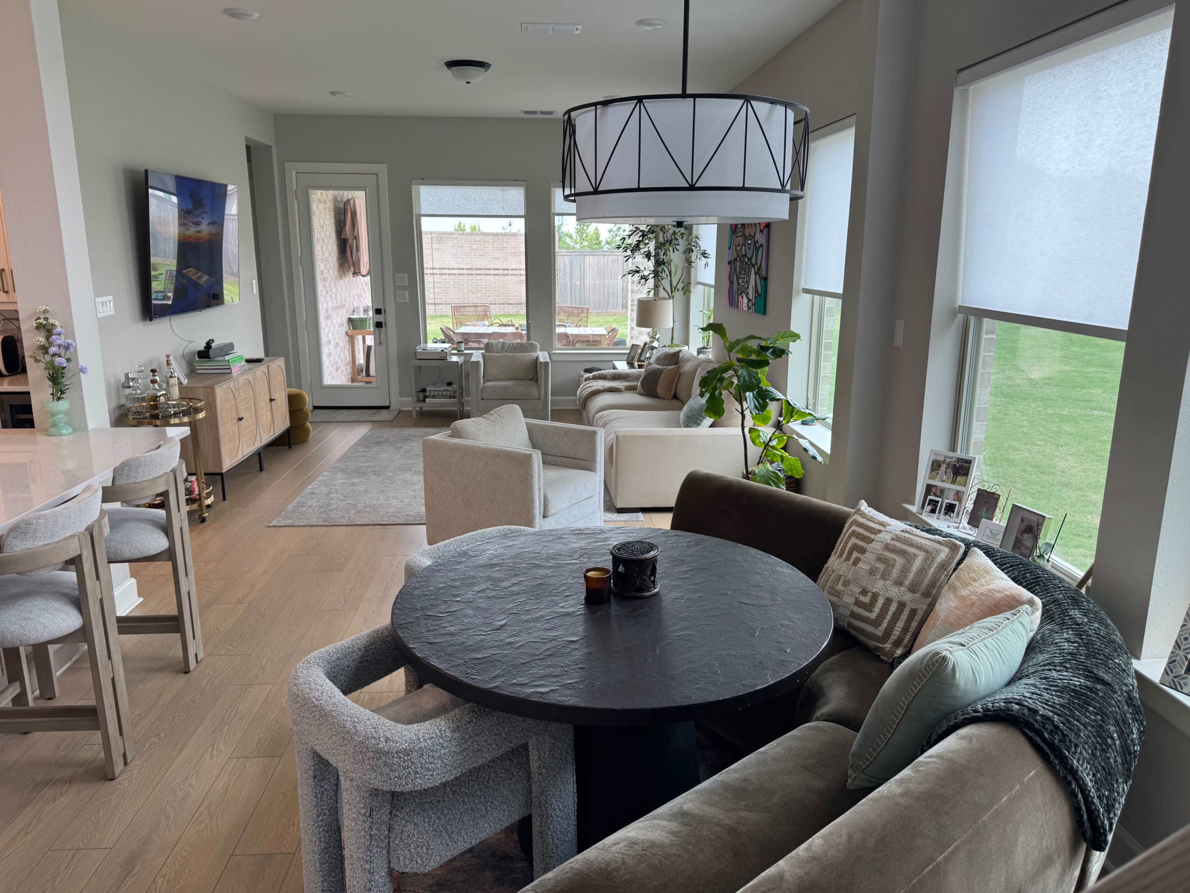 The image shows a modern open-concept living space featuring a dining area with a round black table and a cozy seating area with beige and gray furniture, illuminated by a large chandelier.