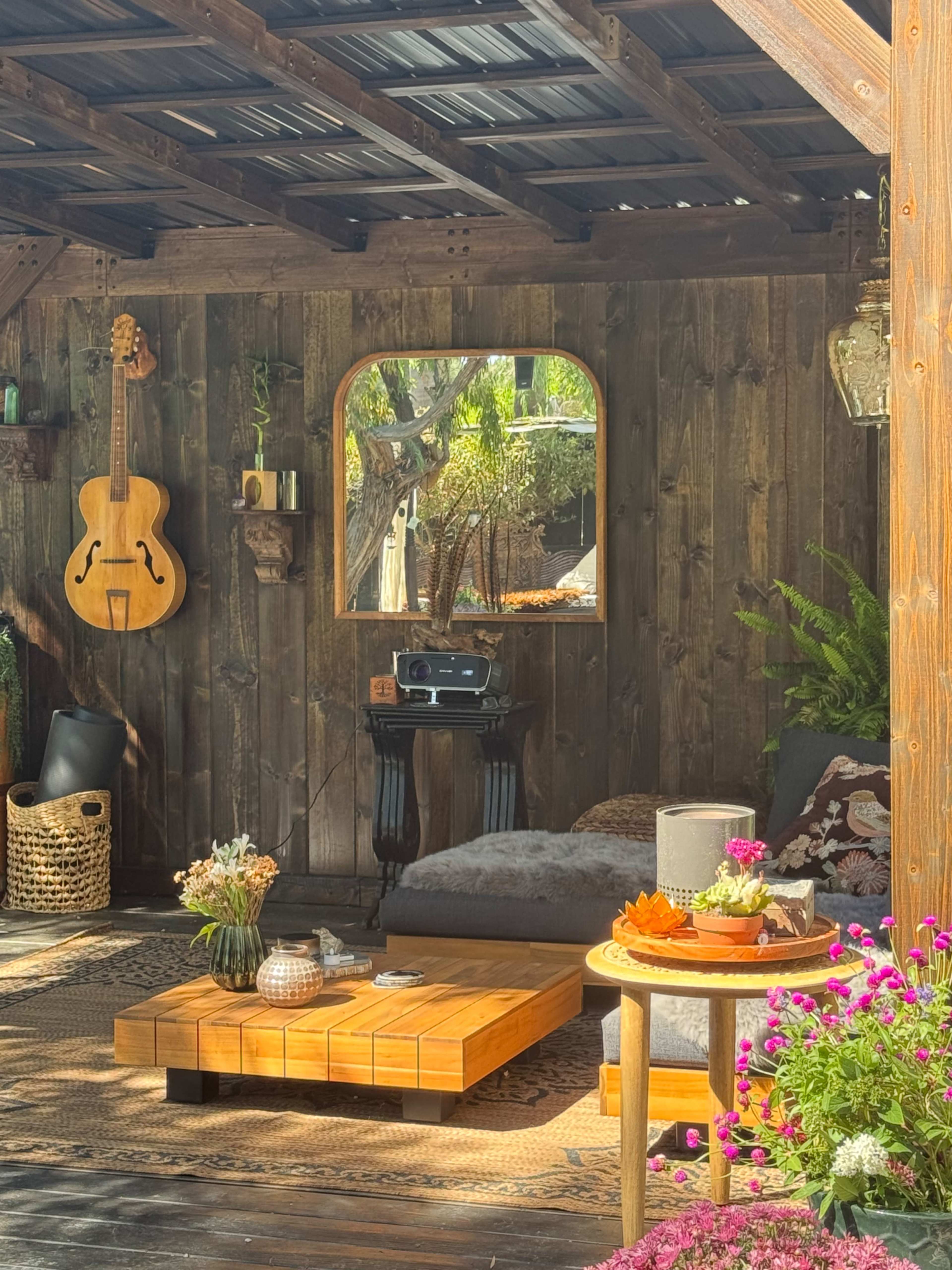 The image shows a cozy outdoor living space featuring a wooden wall, a sofa with decorative pillows, a guitar hanging, a vintage radio on a table, and various plants and flowers arranged around a wooden coffee table.