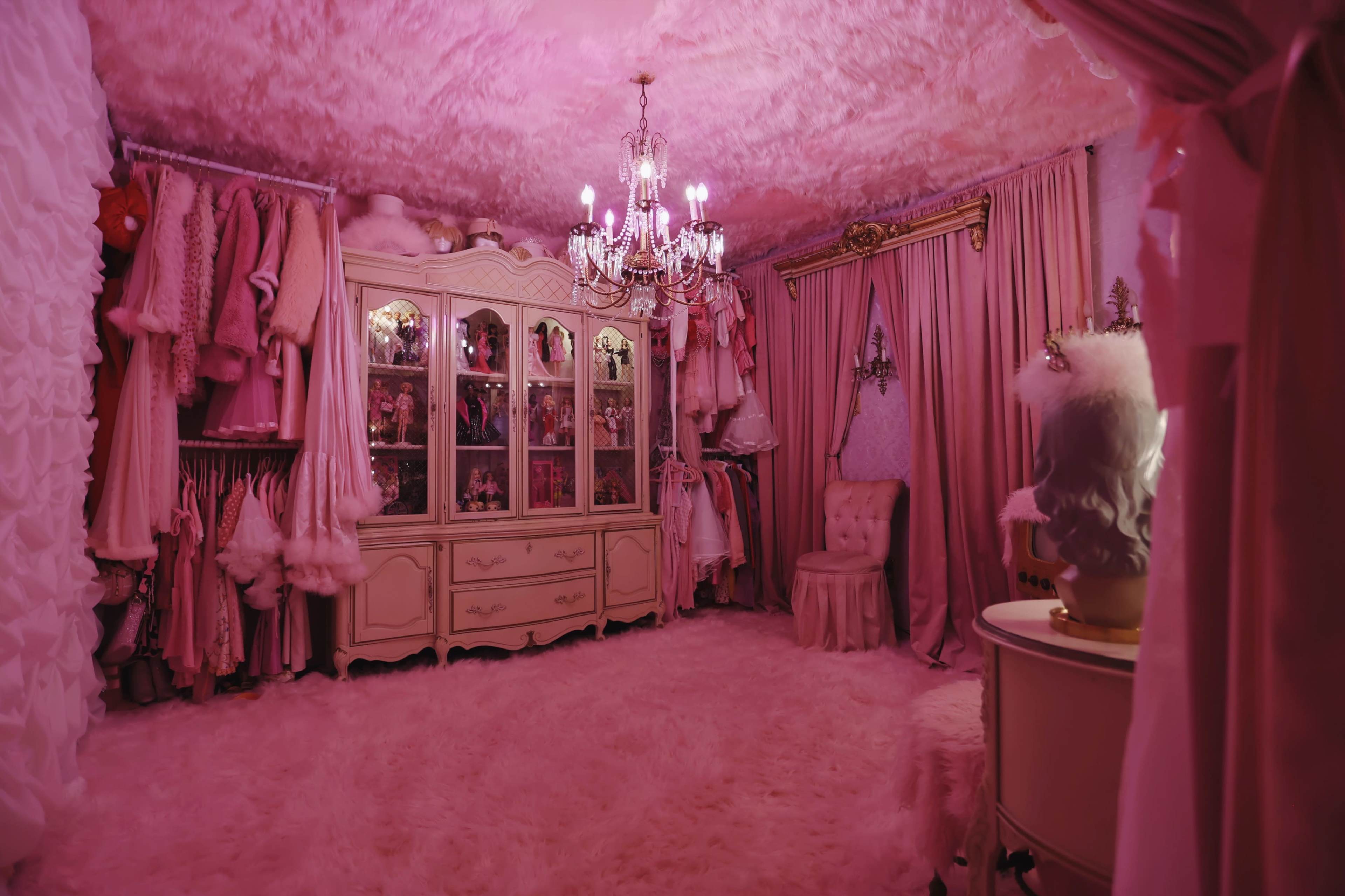 The room features pink walls and decor, a chandelier, and a display cabinet filled with dolls, creating a cozy and whimsical atmosphere.