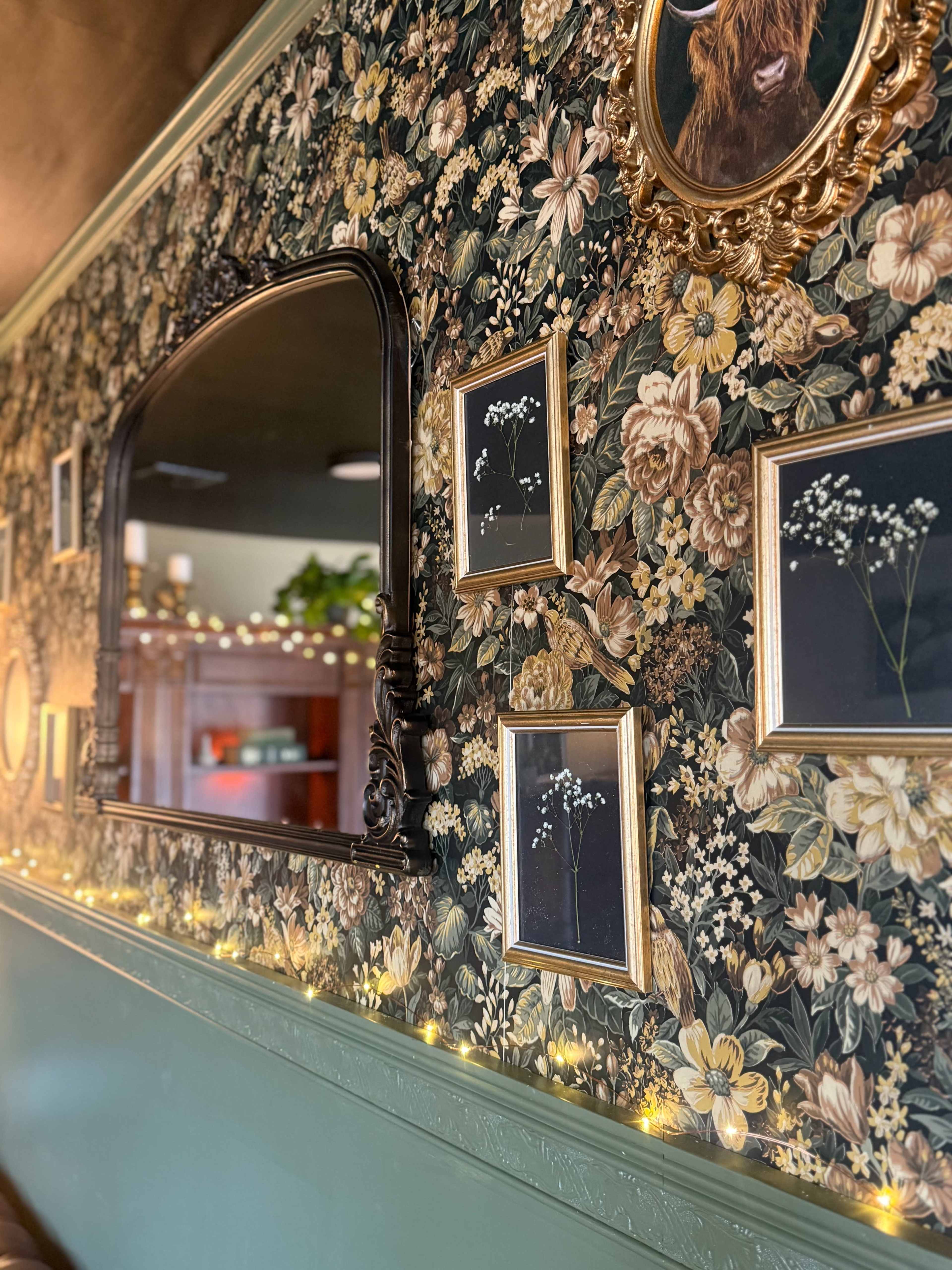 The image shows a wall adorned with floral wallpaper, a mirror, and several framed botanical pictures, illuminated by string lights along the bottom.