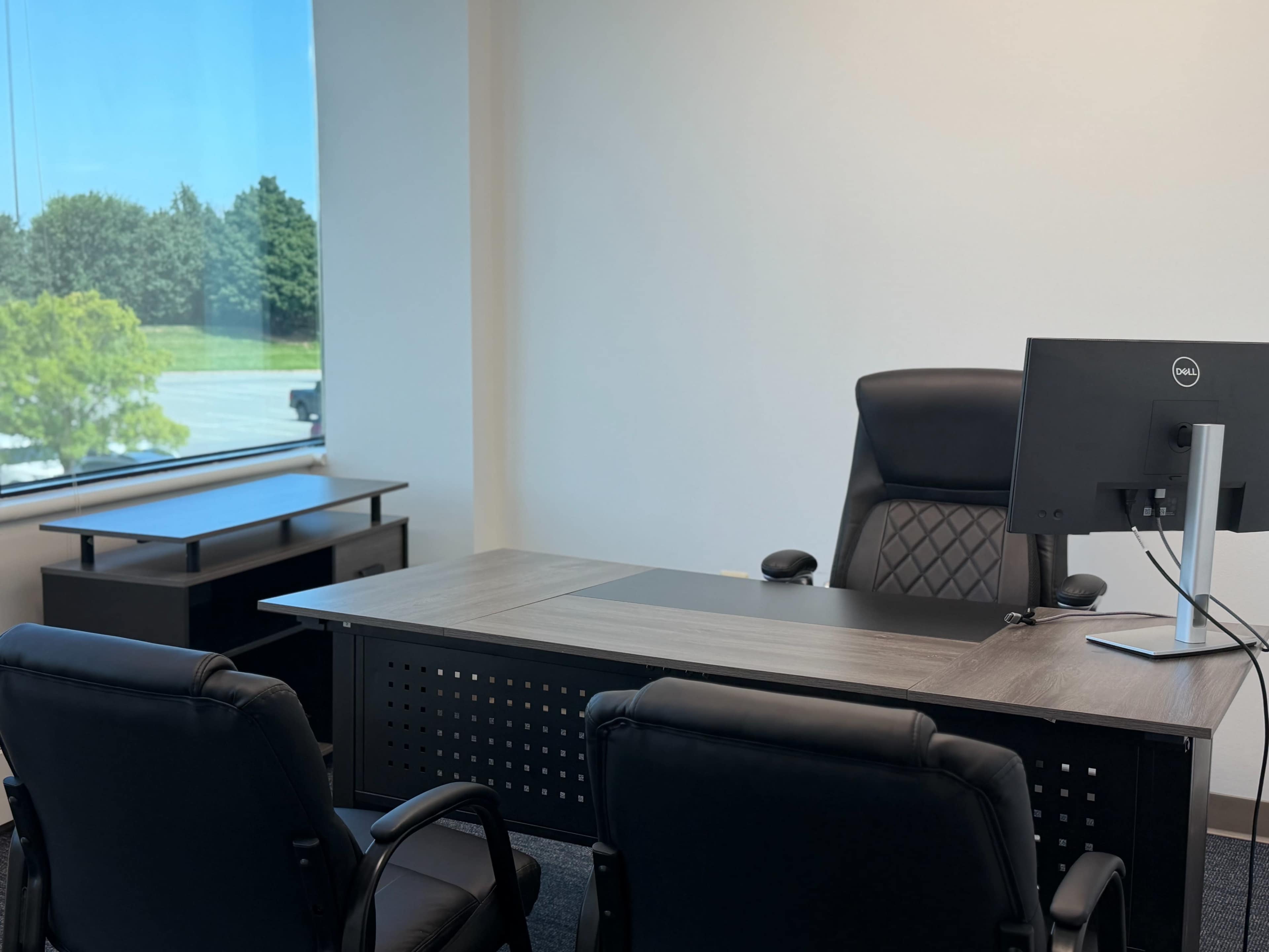 The image shows an empty office with a desk, a computer monitor, and two black chairs facing the desk, set against a large window with trees outside.