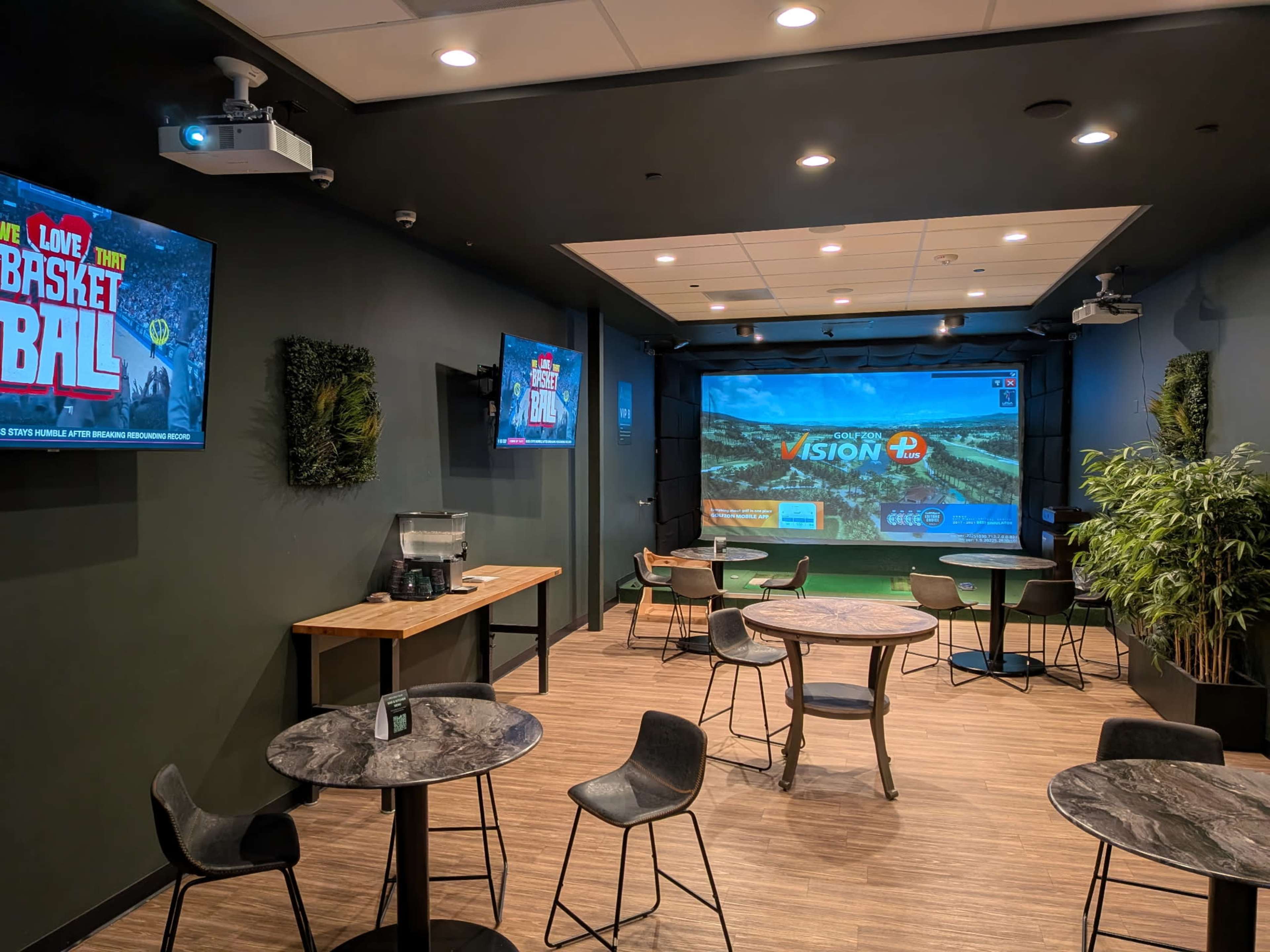 The image depicts a modern indoor space with several small tables and chairs, featuring large screens showing sports content and a projection screen on one wall.