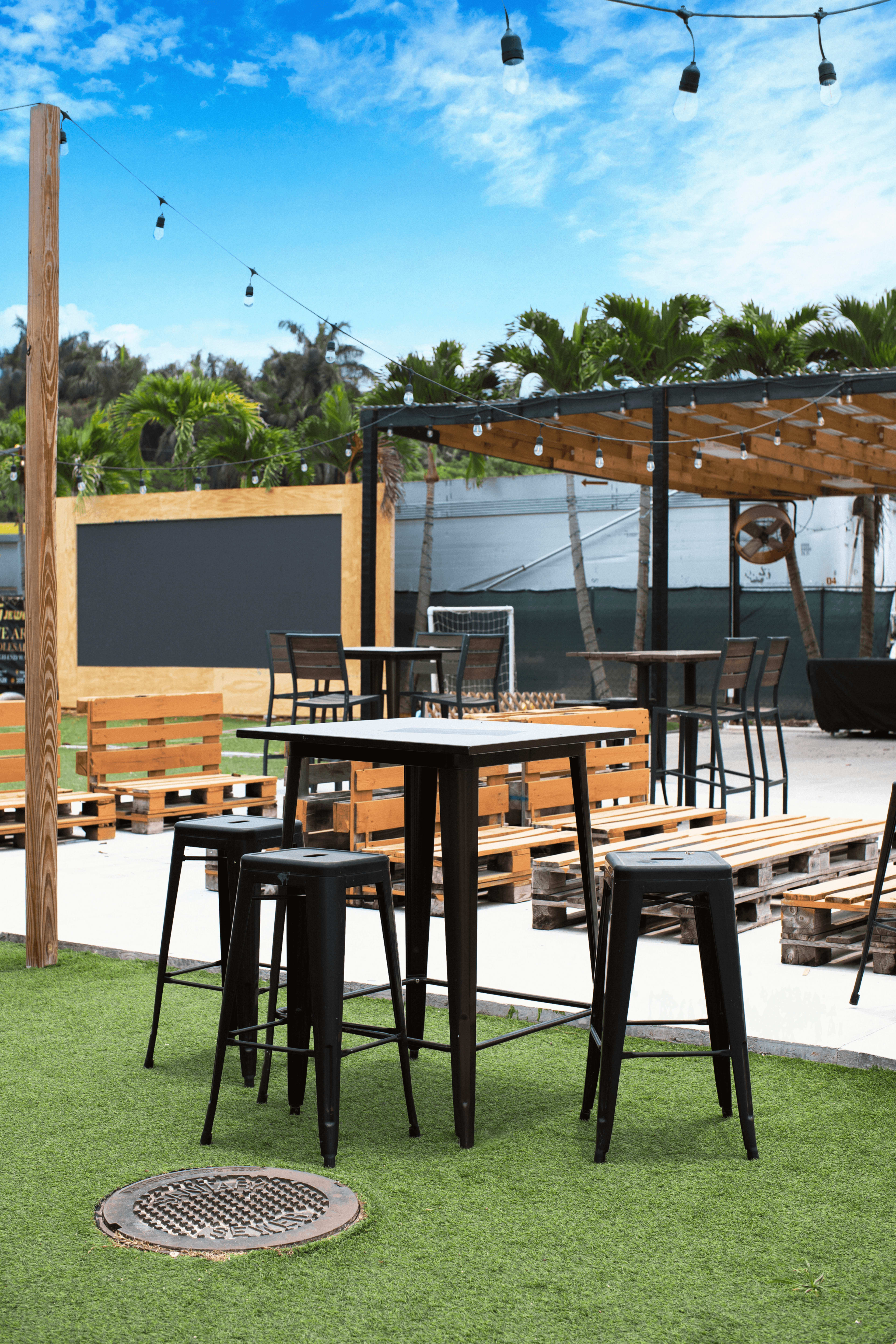 The image shows black metal bar tables and stools arranged on a grassy outdoor area, surrounded by wooden pallets and string lights.