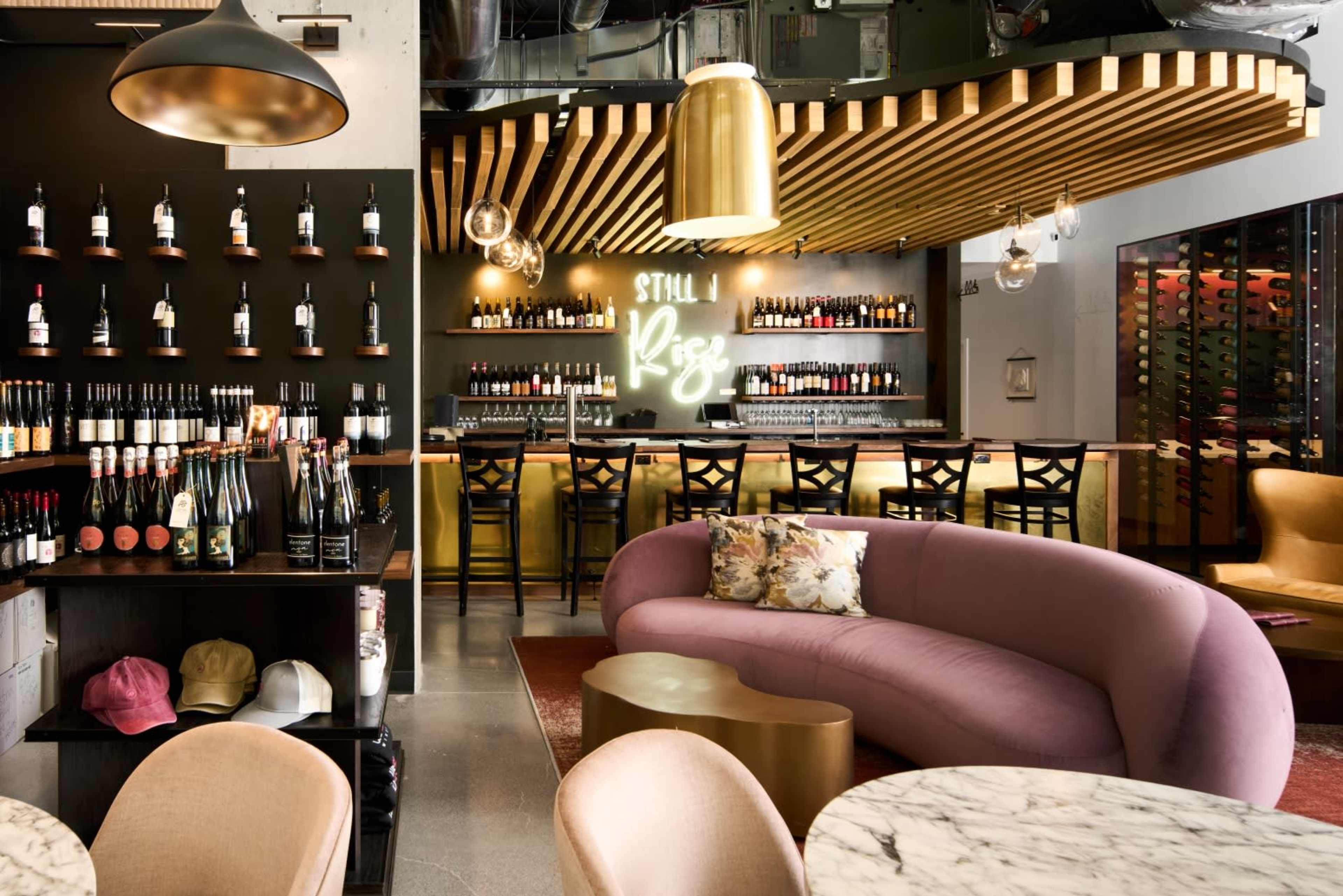 The image shows a stylish bar area featuring a curved purple sofa, a wine bottle display on the wall, and backlit shelving with various bottles behind a sleek counter.