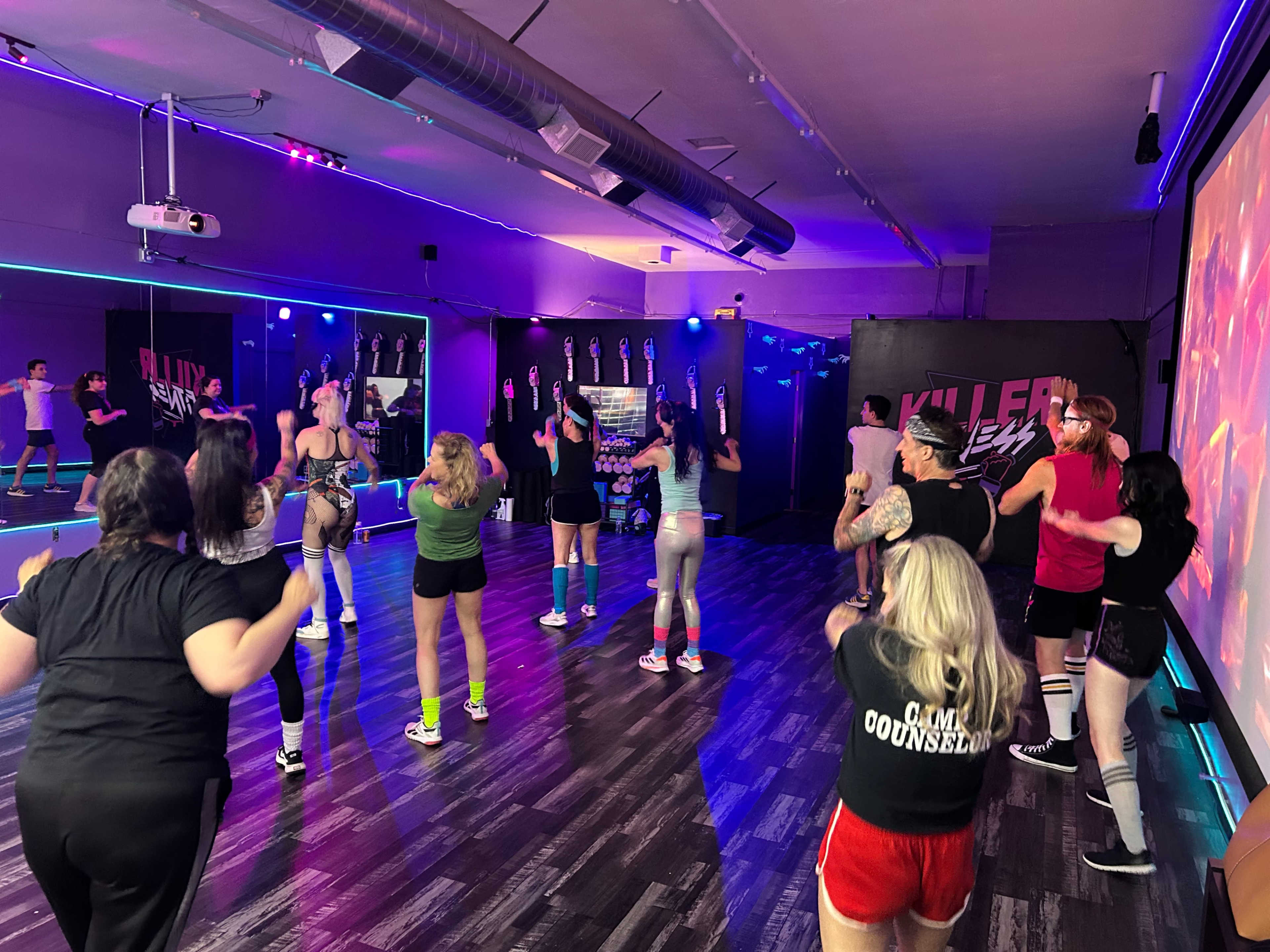 A group of people is participating in a fitness class, following an instructor in a brightly lit studio with colorful lighting and exercise equipment.