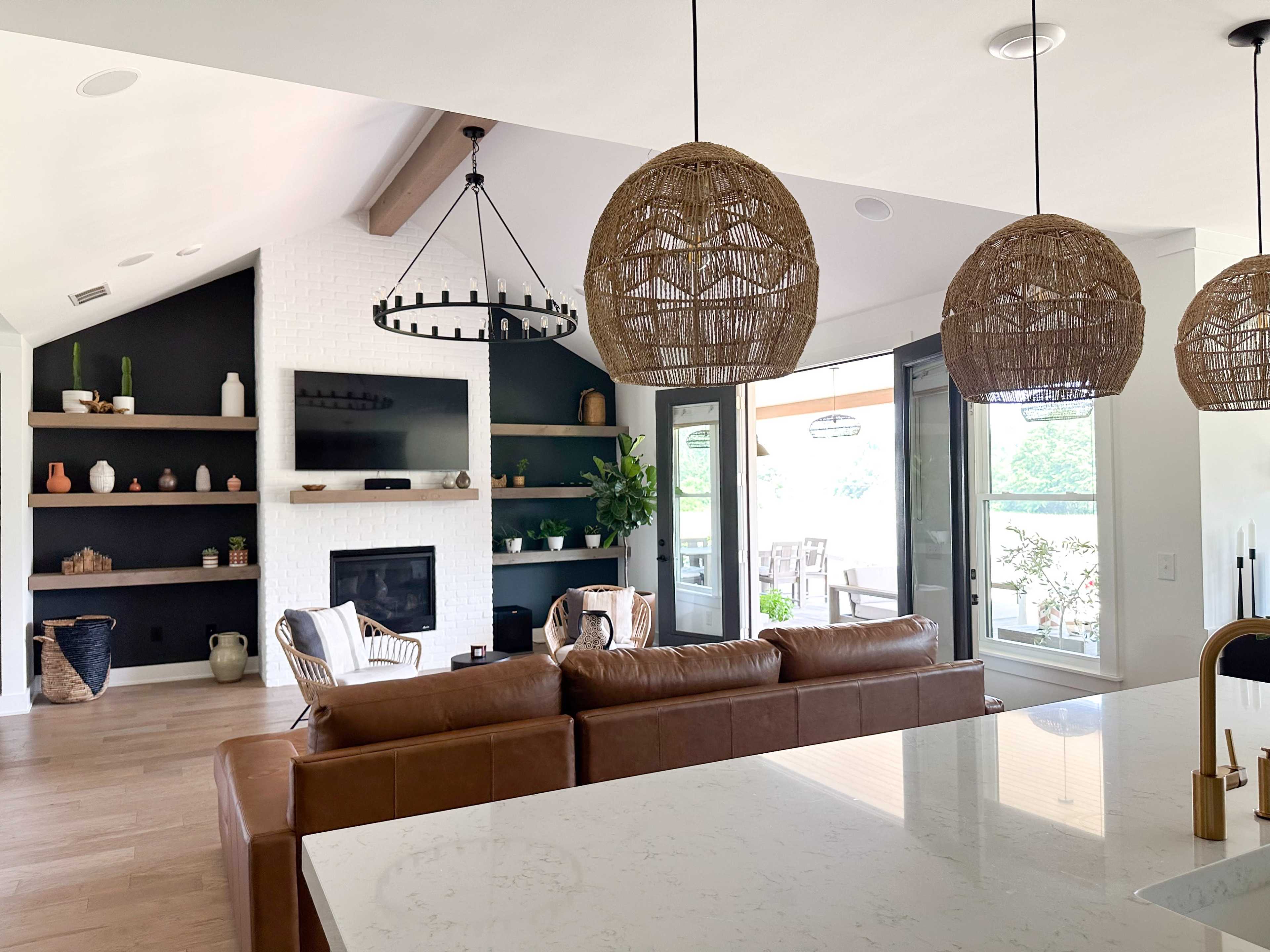 A modern open-concept living area featuring a leather sofa, a television mounted above a fireplace, and a display shelf with decorative items, all illuminated by woven pendant lights.