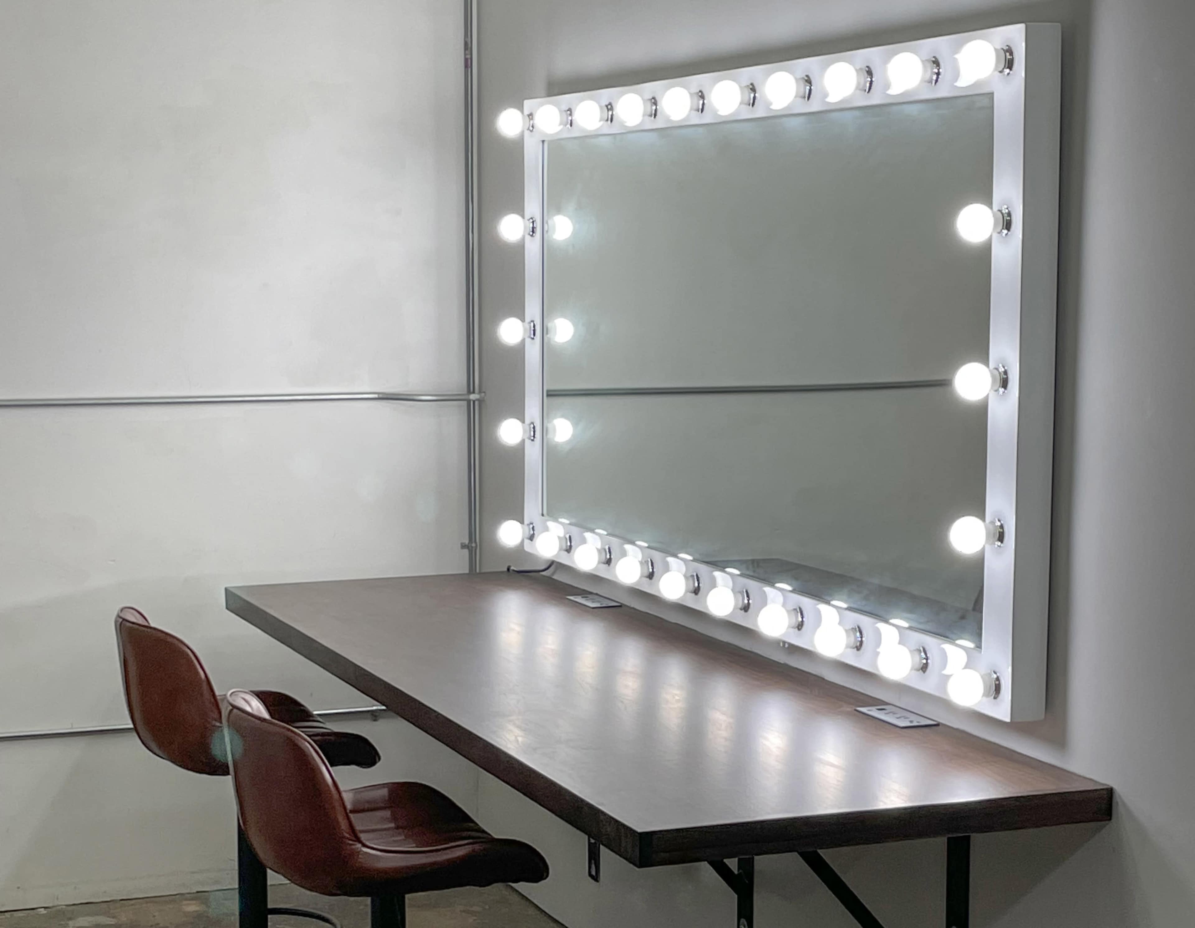 The image shows a large illuminated mirror framed with lights, positioned above a wooden table with two brown chairs.