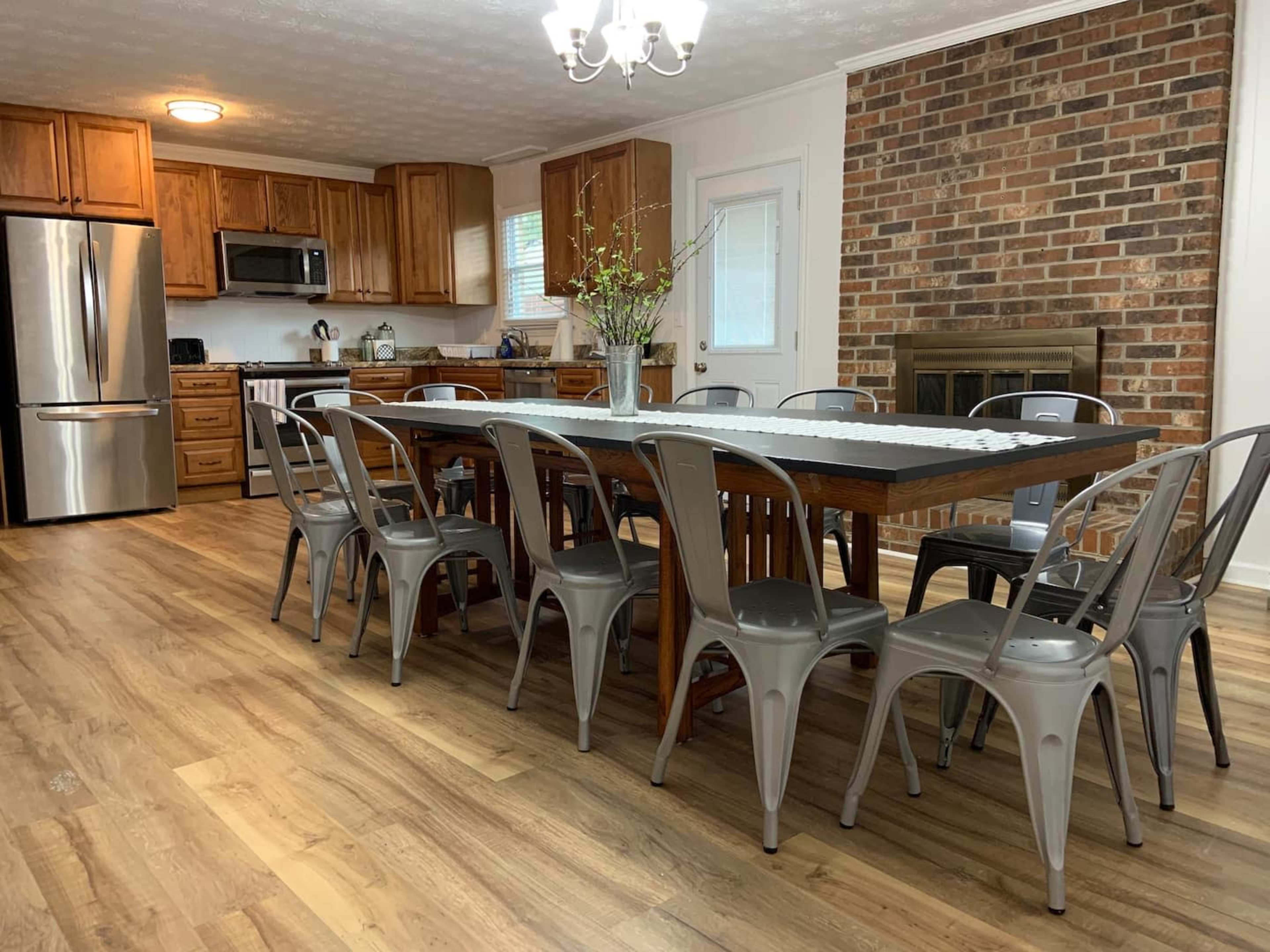 A spacious kitchen and dining area features a large wooden table surrounded by metal chairs, with a brick fireplace and modern appliances in the background.