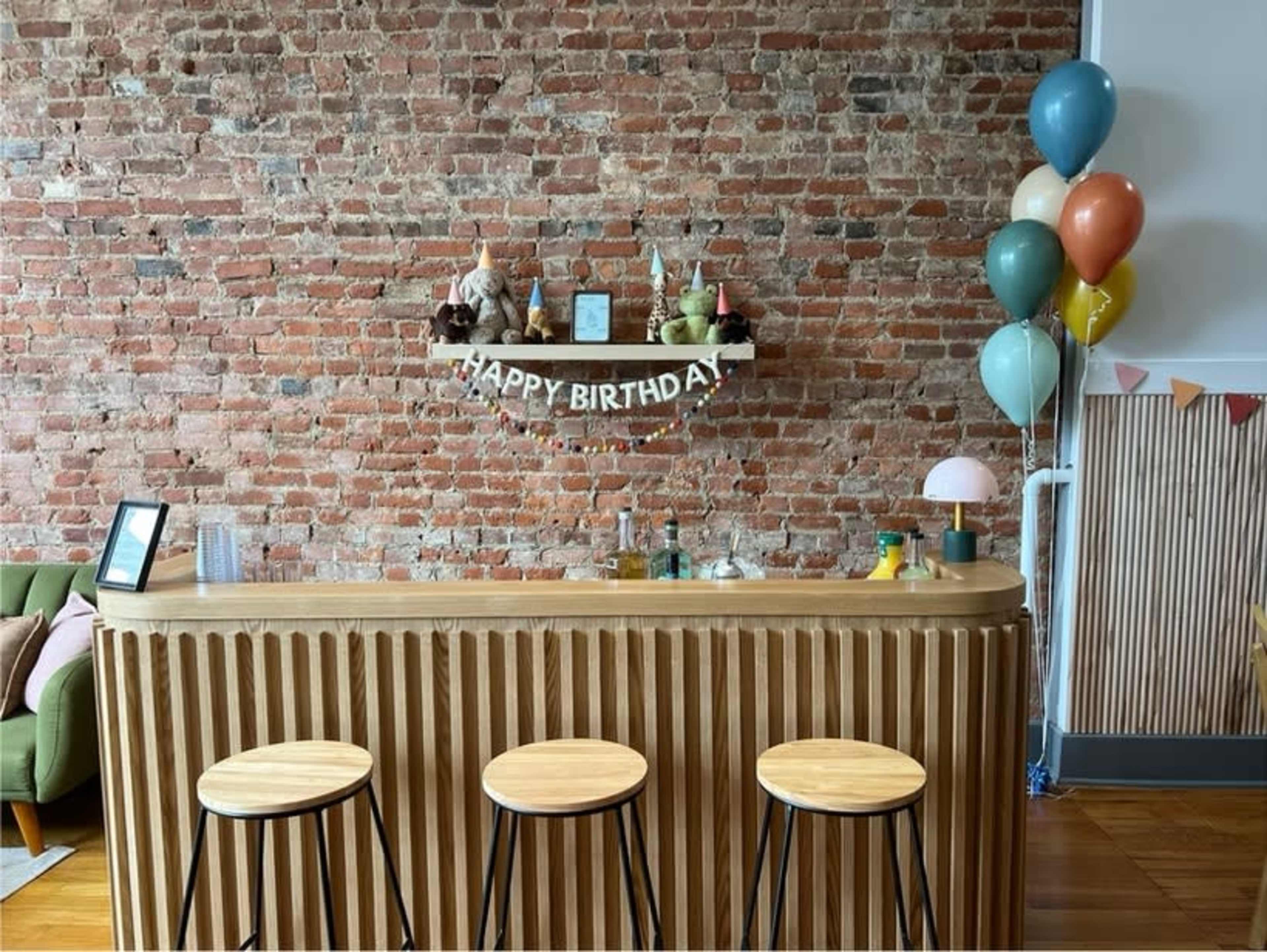 The image shows a wooden bar with three stools in front of a rustic brick wall, adorned with a "Happy Birthday" banner and colorful balloons.