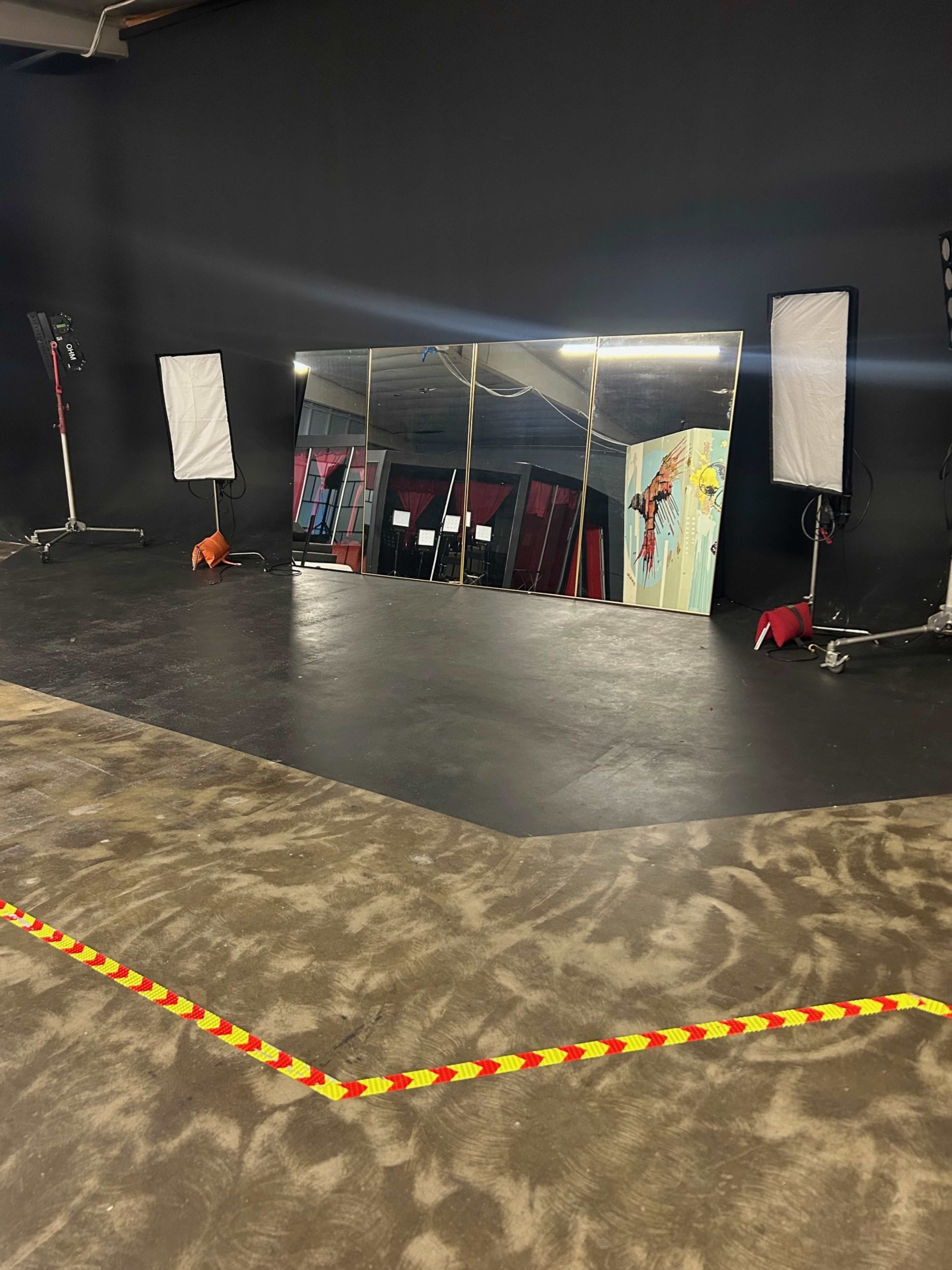 The image shows a studio space with a black wall, a mirrored wall, and lighting equipment on stands, along with a warning tape marking an area on the floor.
