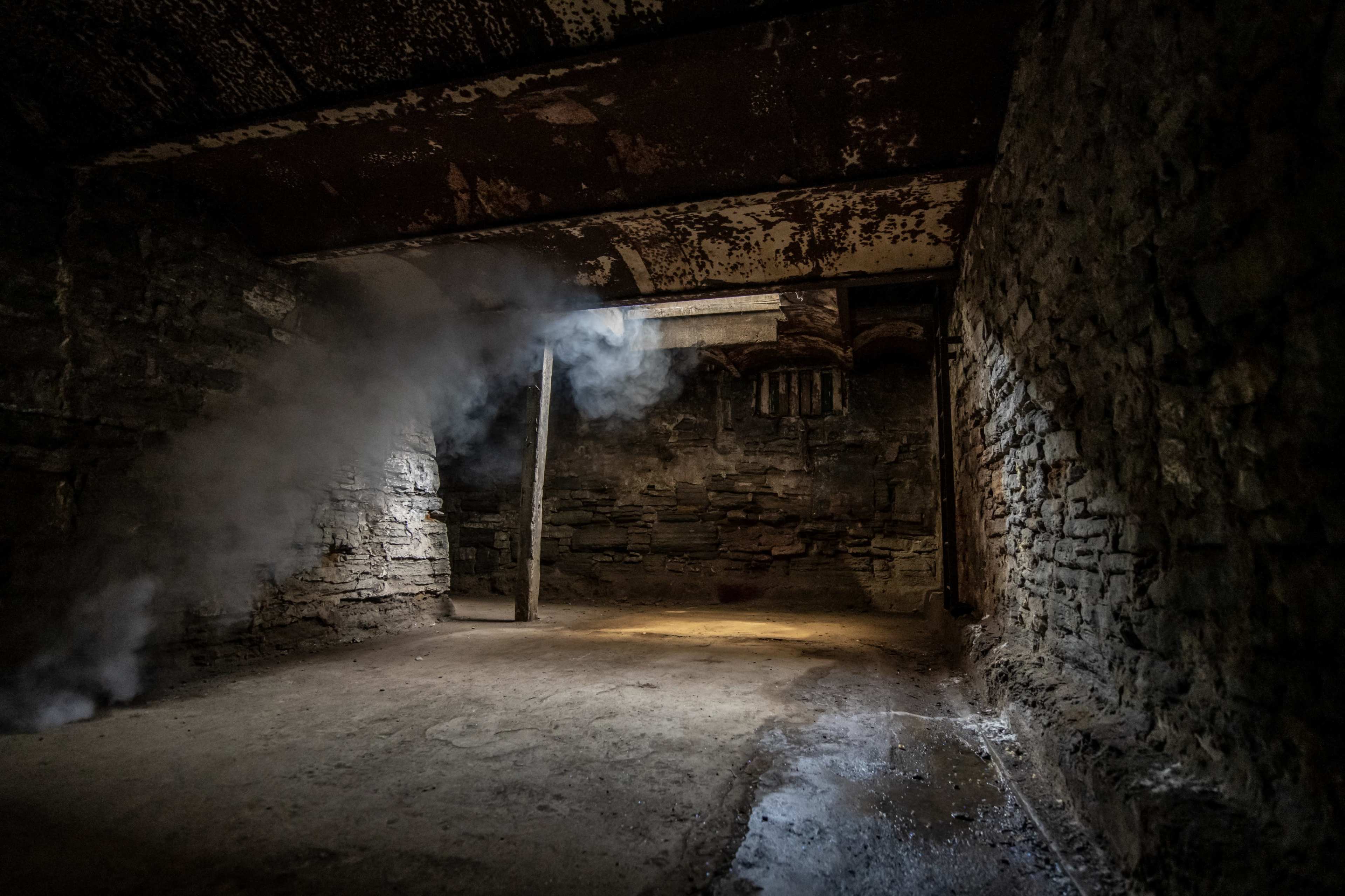 A dimly lit, abandoned room with stone walls, a dirt floor, and smoke curling in the air near a doorway.